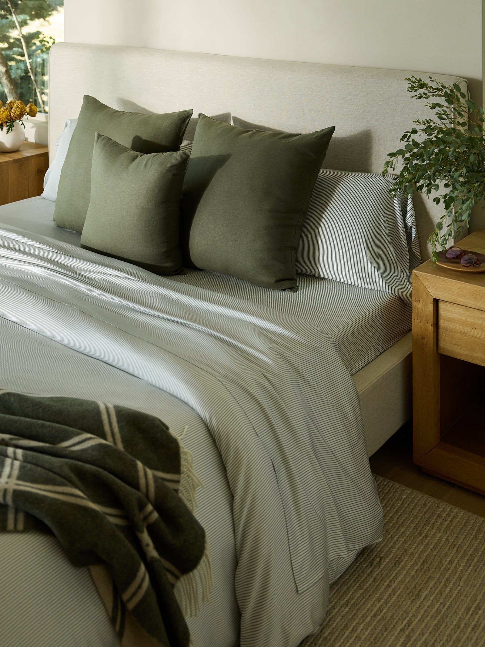 A neatly made bed boasts a light gray headboard and striped bedding, complemented by three green throw pillows. A plaid blanket is elegantly draped at the foot of the bed. Wooden nightstands are positioned on each side, while a plant introduces a touch of greenery to the scene. Enhance your sleep experience with Cozy Earth's Bamboo Flat Sheet for added comfort and style. 