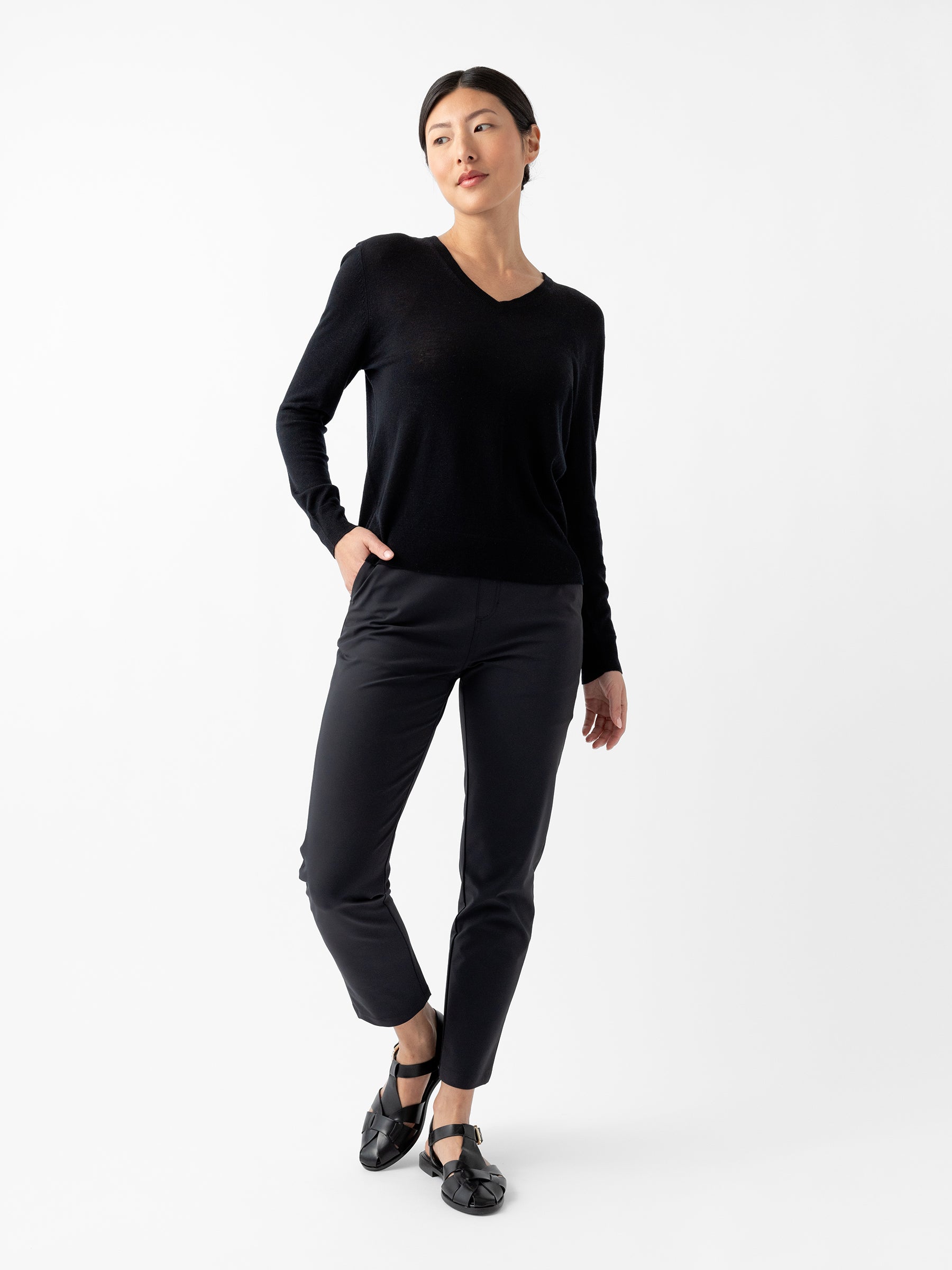 A person stands against a plain white background, dressed in Cozy Earth's Women's AirKnit V-Neck Sweater, black pants, and black shoes. They have one hand in their pocket and gaze slightly off-camera. The overall look is minimalist and chic. |Color:Jet Black