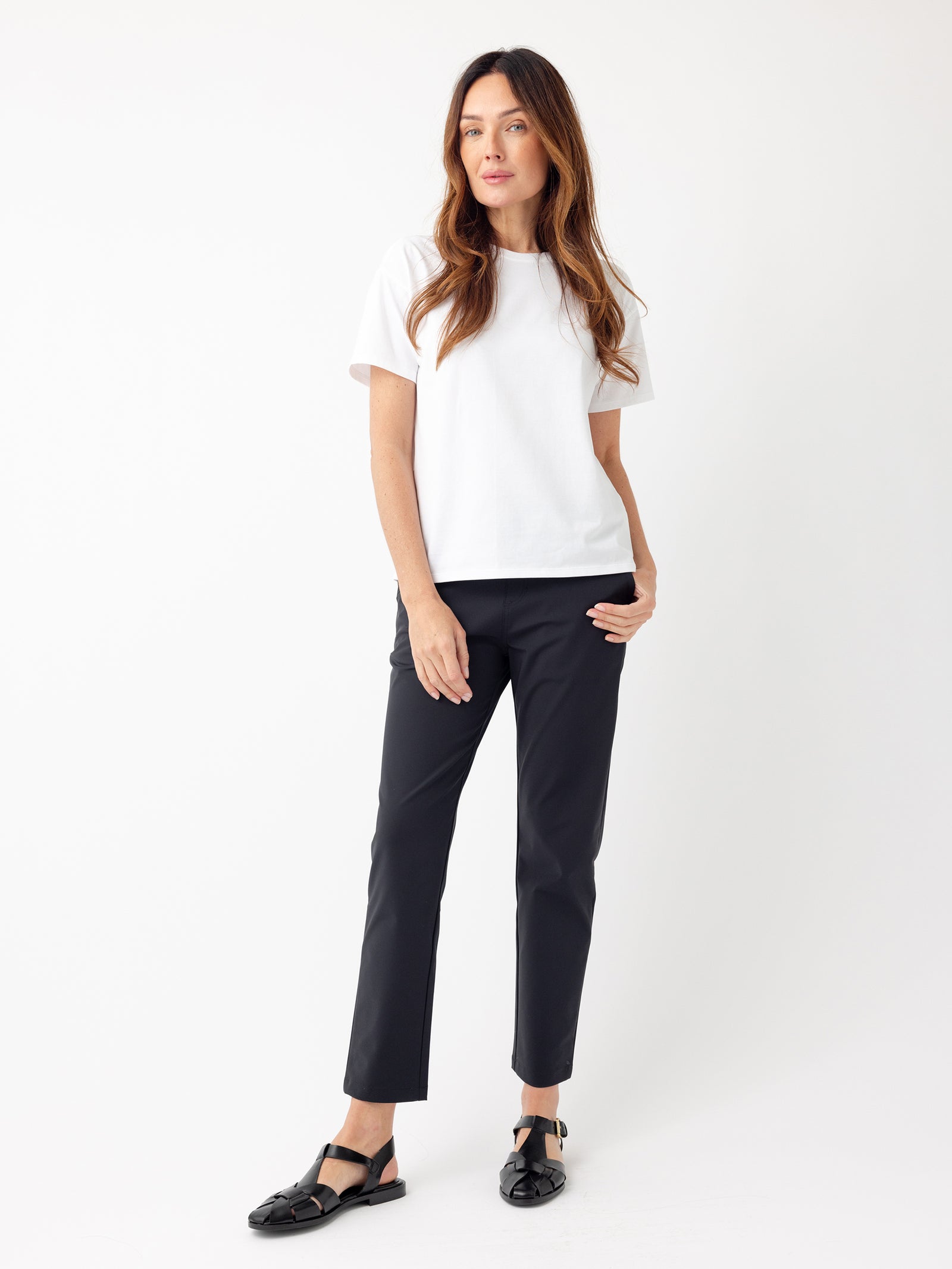 Against a plain white backdrop stands a woman with long, wavy brown hair. She is dressed in a white short-sleeve t-shirt and the Women's Always Cropped Pant from Cozy Earth in black. Completing her outfit are black flat shoes. Her right hand rests on her hip, and she gazes slightly to the left. 