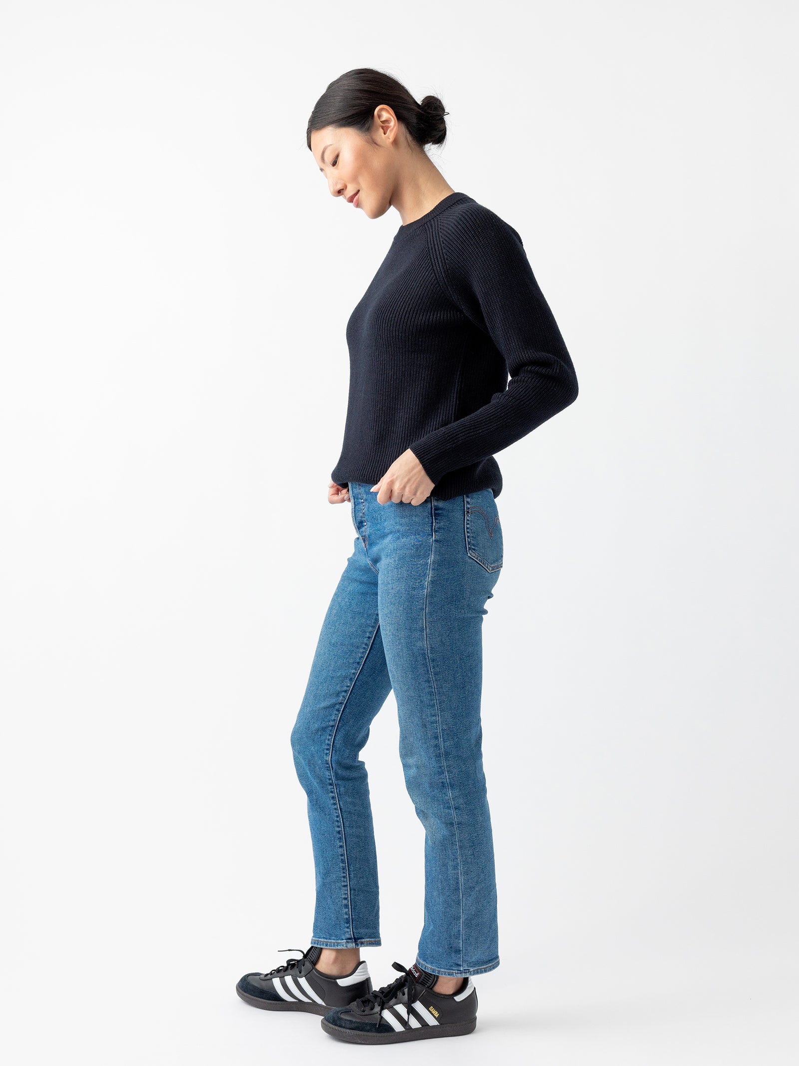 A woman wearing a Cozy Earth Women's Classic Crewneck, blue jeans, and black sneakers with white stripes stands against a plain white background. She is facing slightly to the left with her hands in her pockets and her hair tied back. 