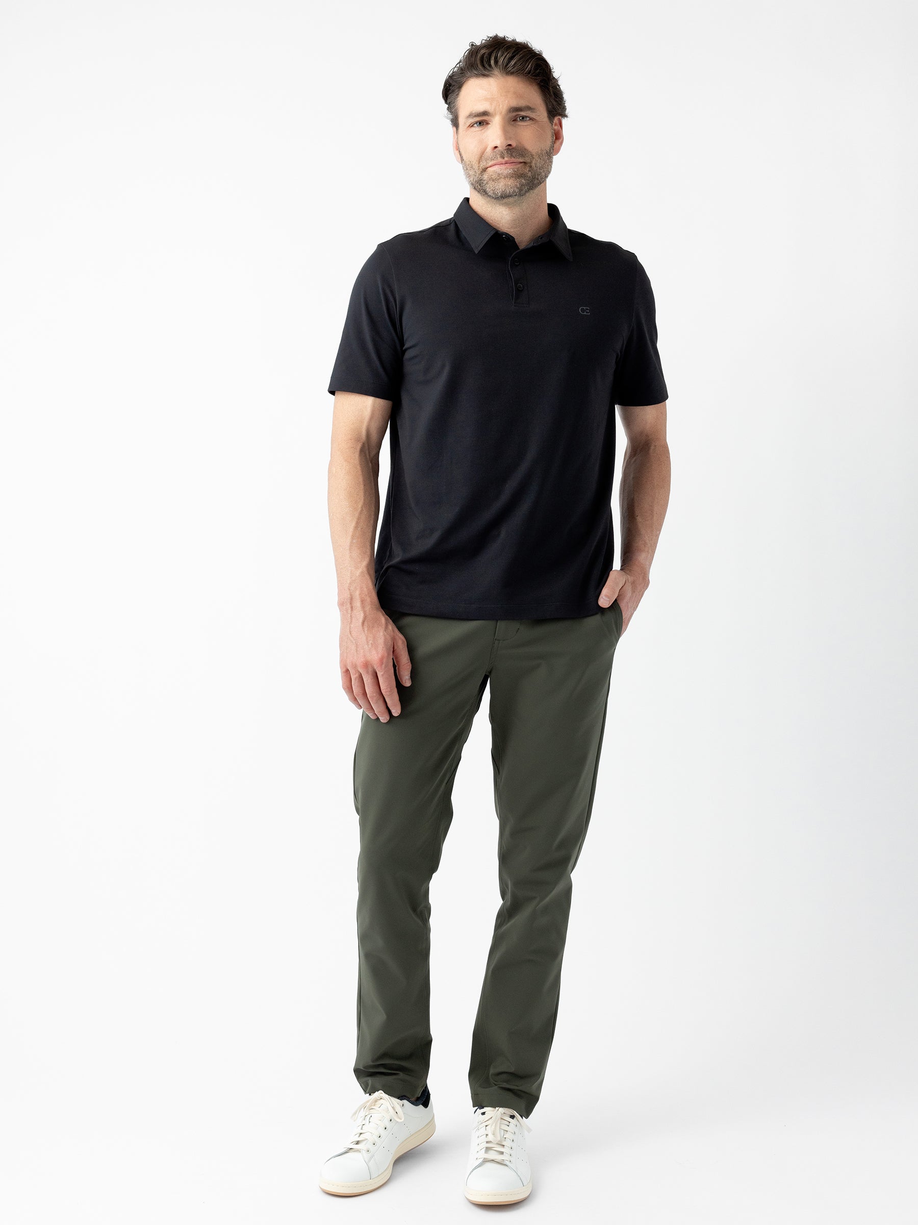 A man with short dark hair and a beard stands against a white background. He is wearing Cozy Earth's Men's Everyday Polo in black, olive green pants, and white sneakers. His left hand is in his pants pocket, and he has a relaxed expression. |Color:Jet Black