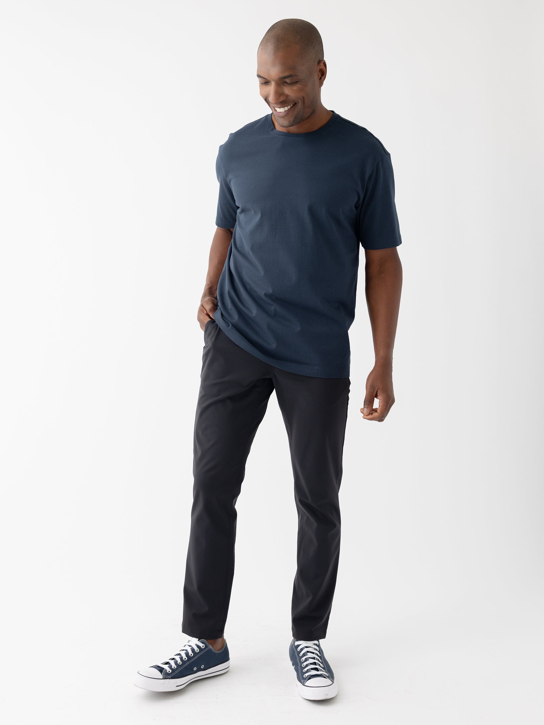 A smiling man wears a navy blue t-shirt, Cozy Earth's Men's Everywhere Pant 30L in black, and navy blue sneakers. He stands against a plain white background with his left hand in his pocket and his right hand resting at his side. |Color:Jet Black