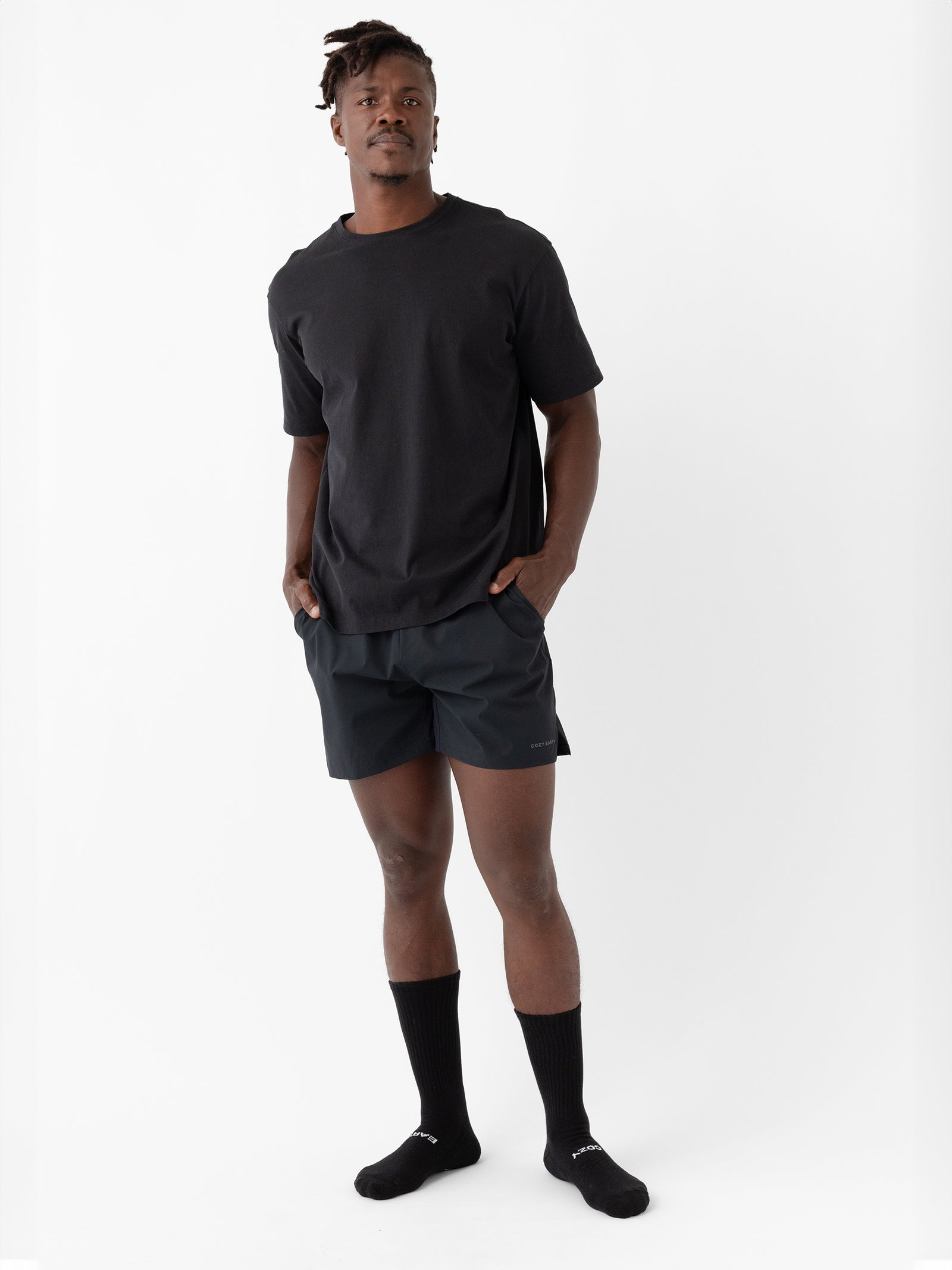 A man stands against a plain white background, wearing a black t-shirt, Cozy Earth's Men's Performance Sleep Shorts in black, black socks, and black shoes. His hands are in the pockets of his shorts, and he has a neutral expression on his face. 