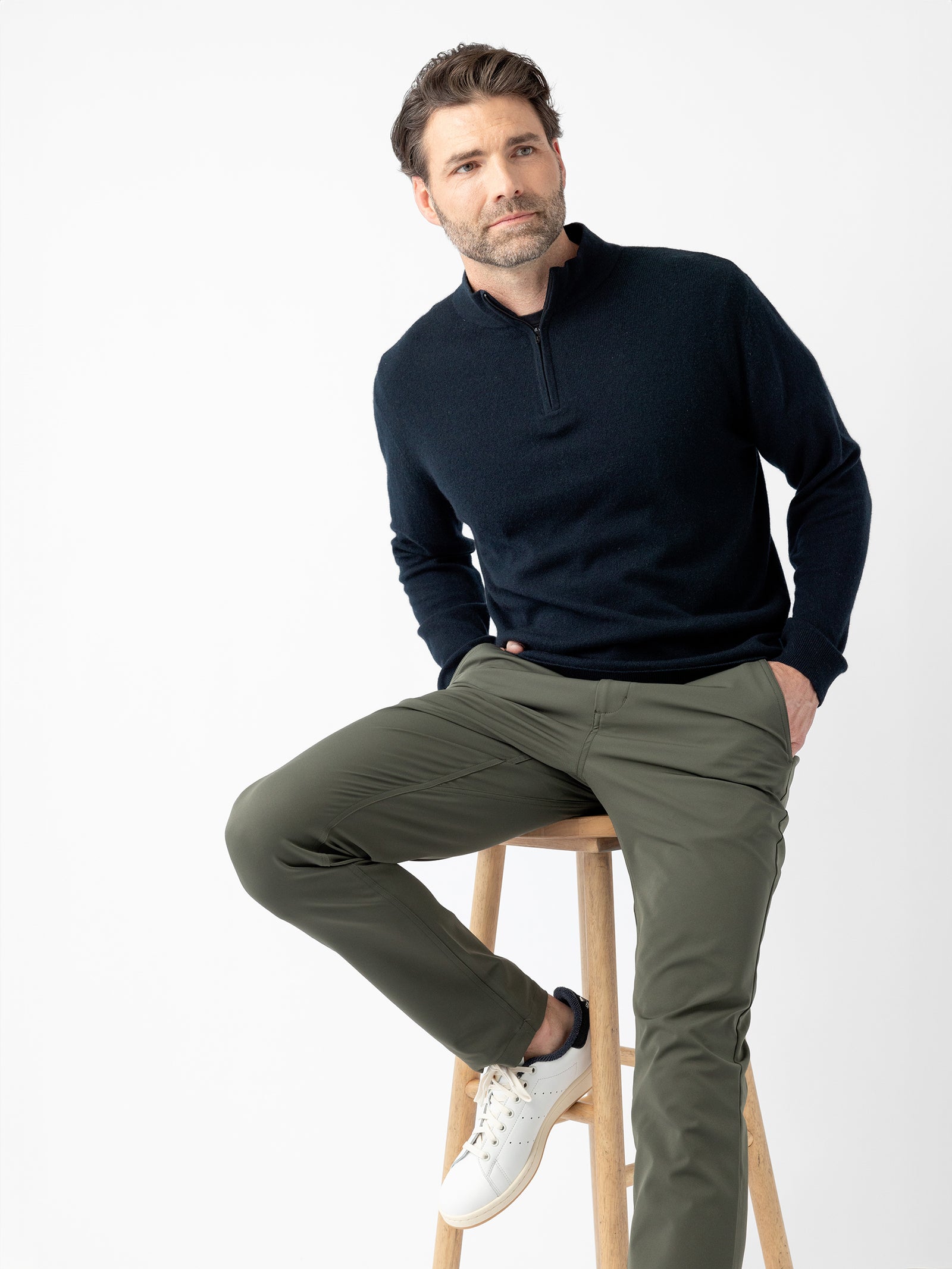 A man with short dark hair and a beard is seated on a wooden stool. He is wearing a navy blue Men's Quarter Zip Sweater by Cozy Earth, olive green pants, and white sneakers. He has his hands casually in his pockets and is looking off to the side against a plain white background. 