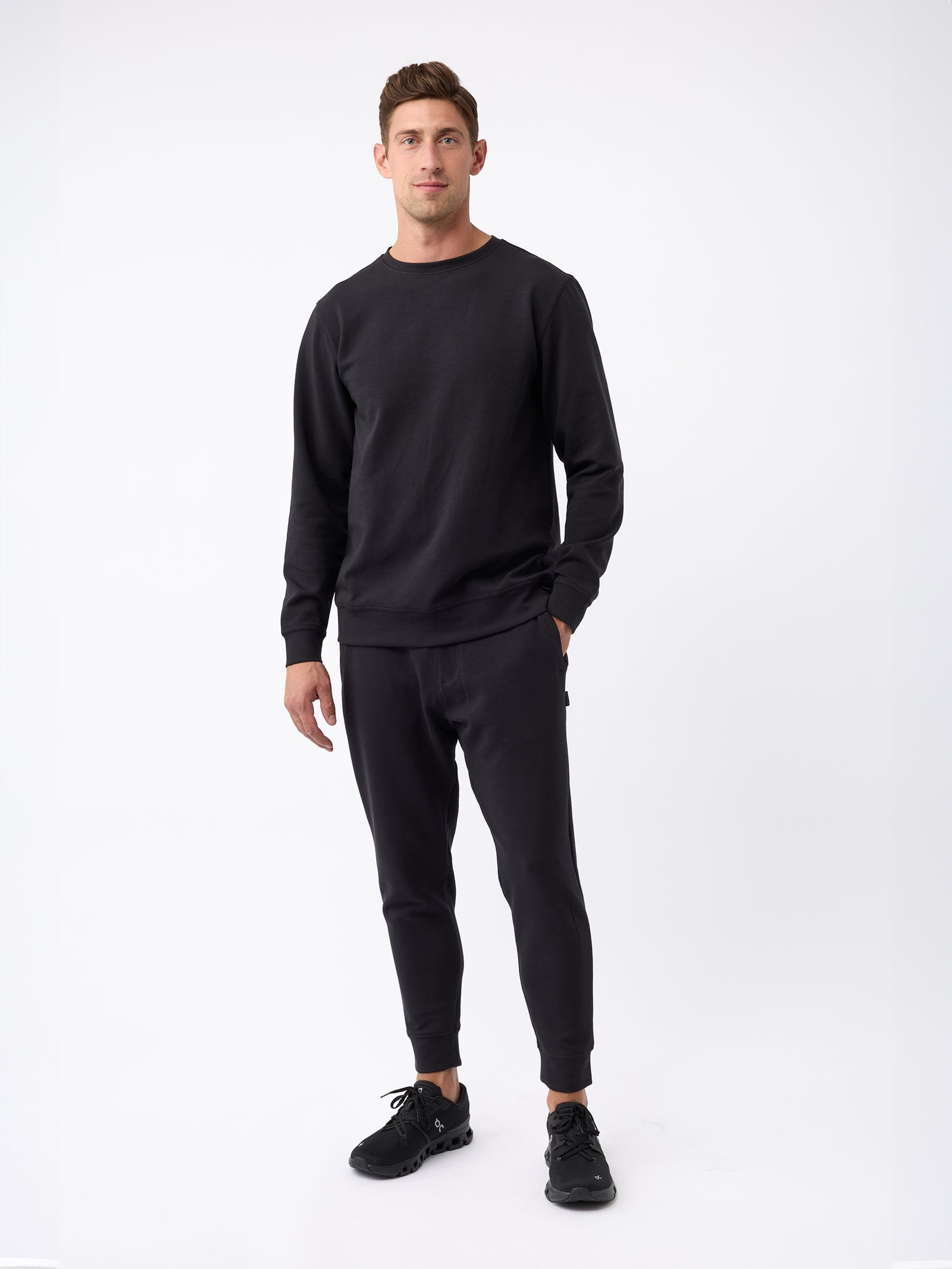 A man stands against a plain white background, wearing the Cozy Earth Men's StretchTech Crewneck along with black pants and black sneakers. He has one hand in his pocket and looks directly at the camera. 