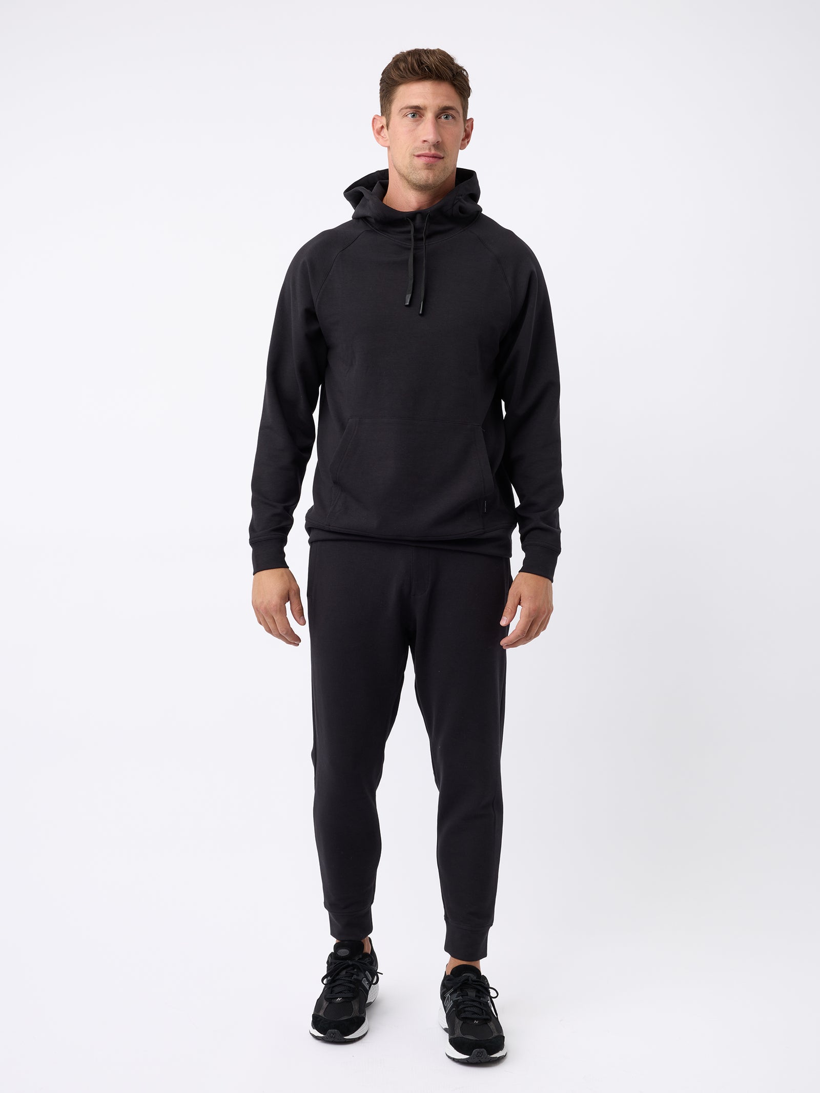 A person stands against a plain white background wearing a Cozy Earth Men's StretchTech Hoodie and black jogger pants. They have short, brown hair and are looking forward with a neutral expression. They are also wearing black athletic shoes. 