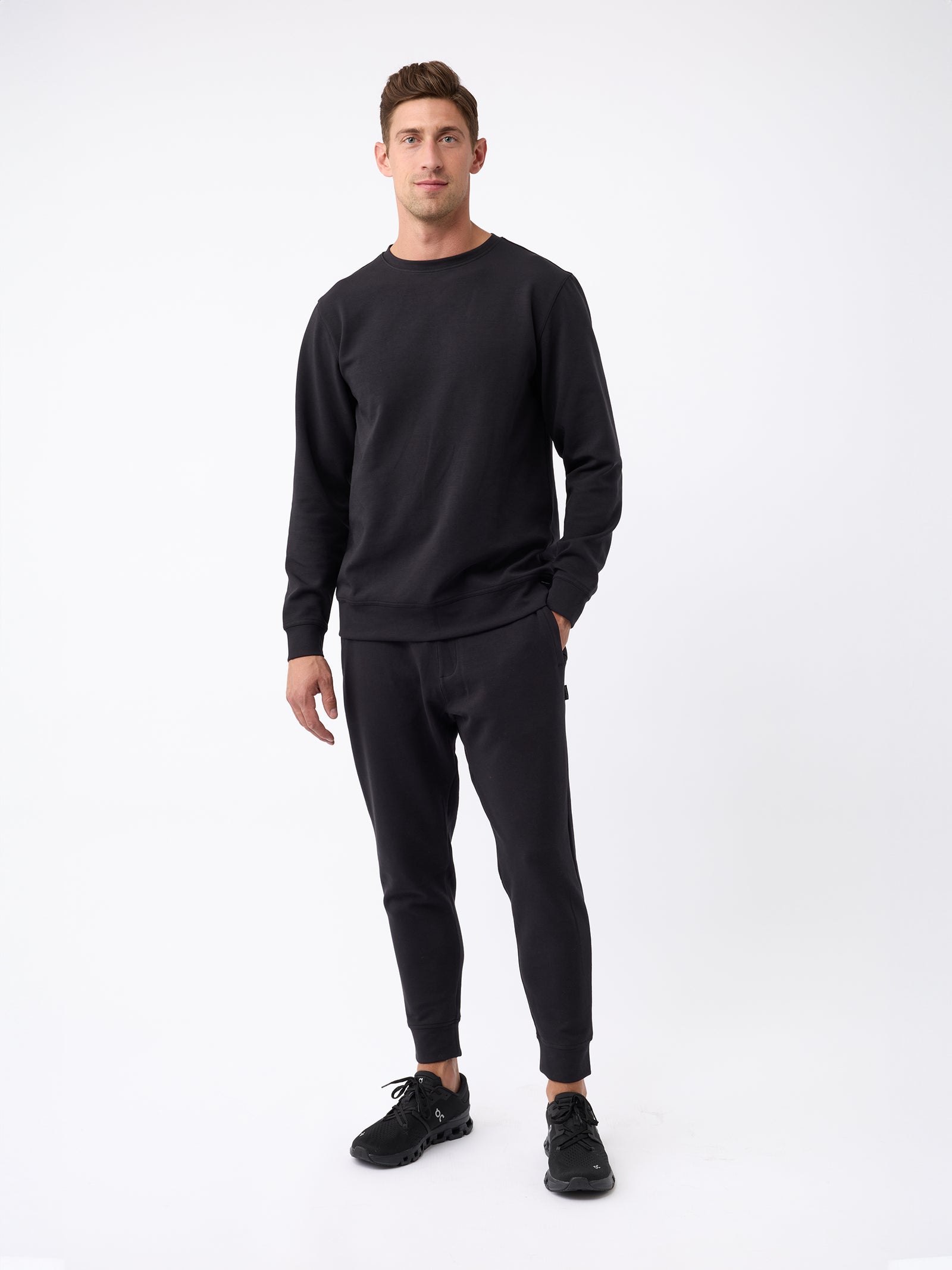 A man stands against a plain white background wearing a black Men's StretchTech Jogger from Cozy Earth. He has one hand in his pocket, is also wearing black shoes, and is looking straight ahead with a relaxed expression. 