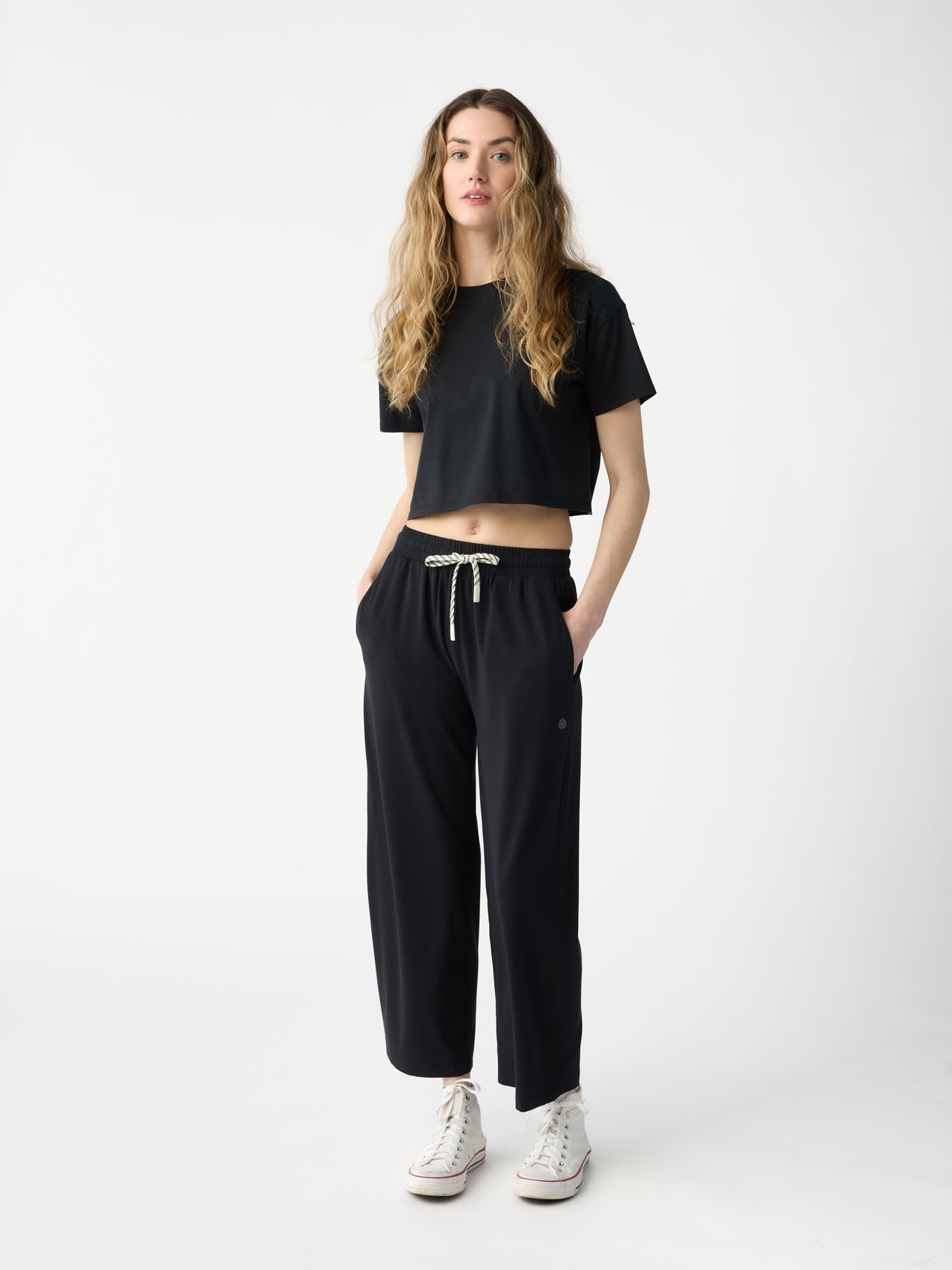 A woman with long wavy hair wears a black cropped t-shirt, Women's Studio Cropped Wide Leg Pant by Cozy Earth in black, and white sneakers. She stands with her hands in her pockets against a plain white background. 