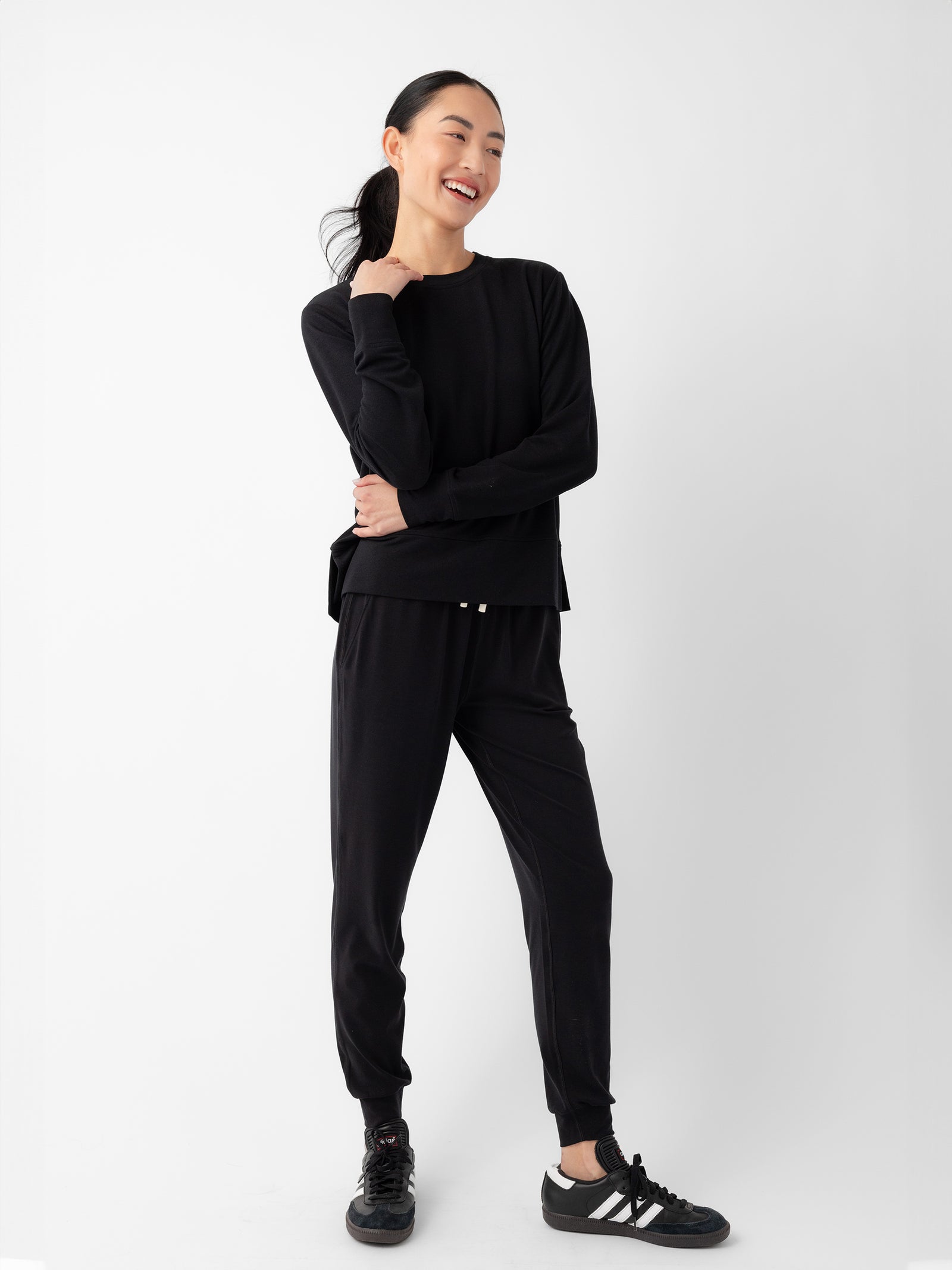 A woman stands against a plain white background, smiling and looking to her left. She is dressed in a black Women's StudioLite Crewneck by Cozy Earth and matching jogger pants with black and white athletic shoes. She has her dark hair tied back in a ponytail and one hand resting on her chin. 