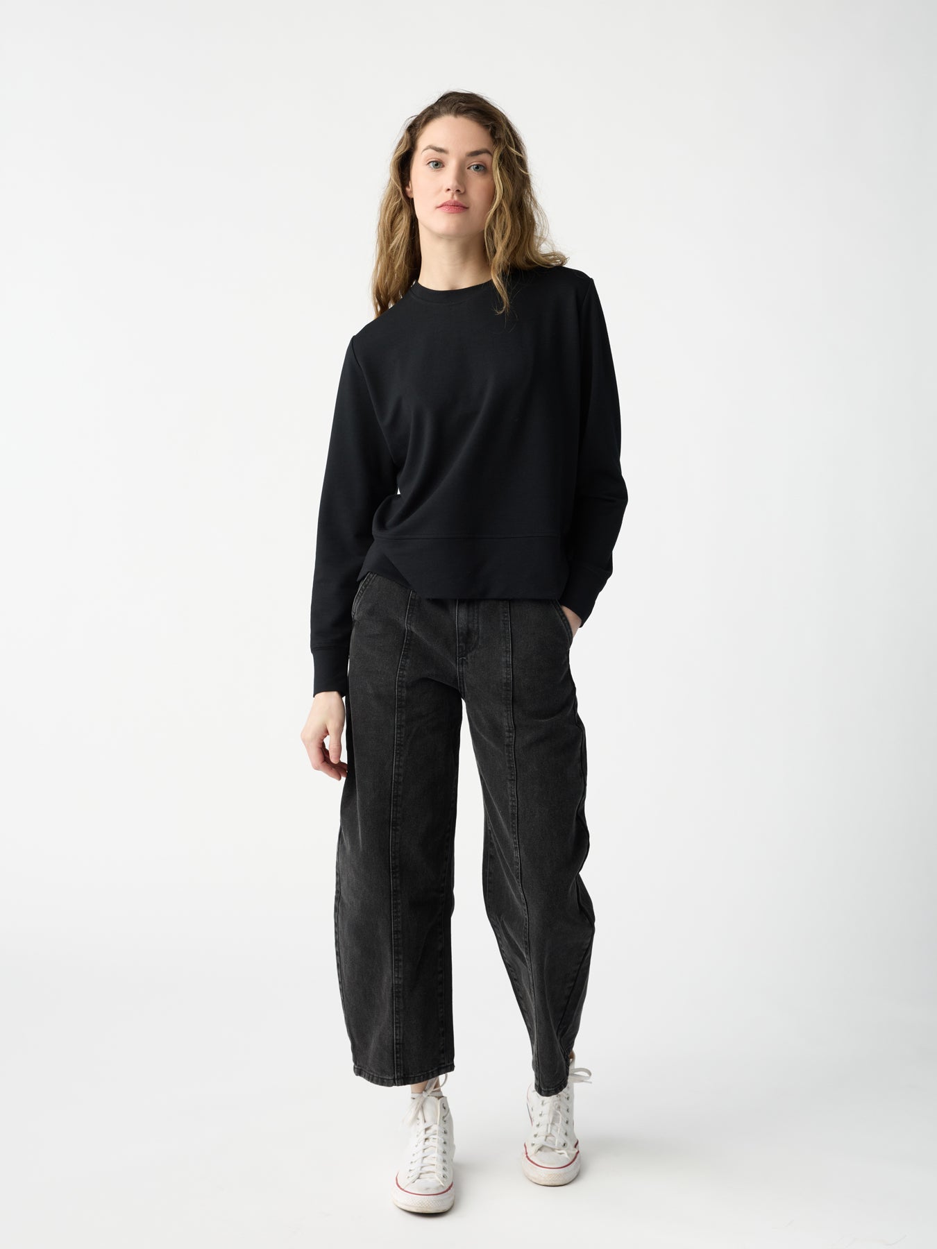 Wearing the black Women's StudioLite Crewneck by Cozy Earth, paired with dark wide-leg jeans and white high-top sneakers, a person stands against a plain white background. With long wavy hair and facing the camera, they maintain a relaxed expression. |Color:Jet Black