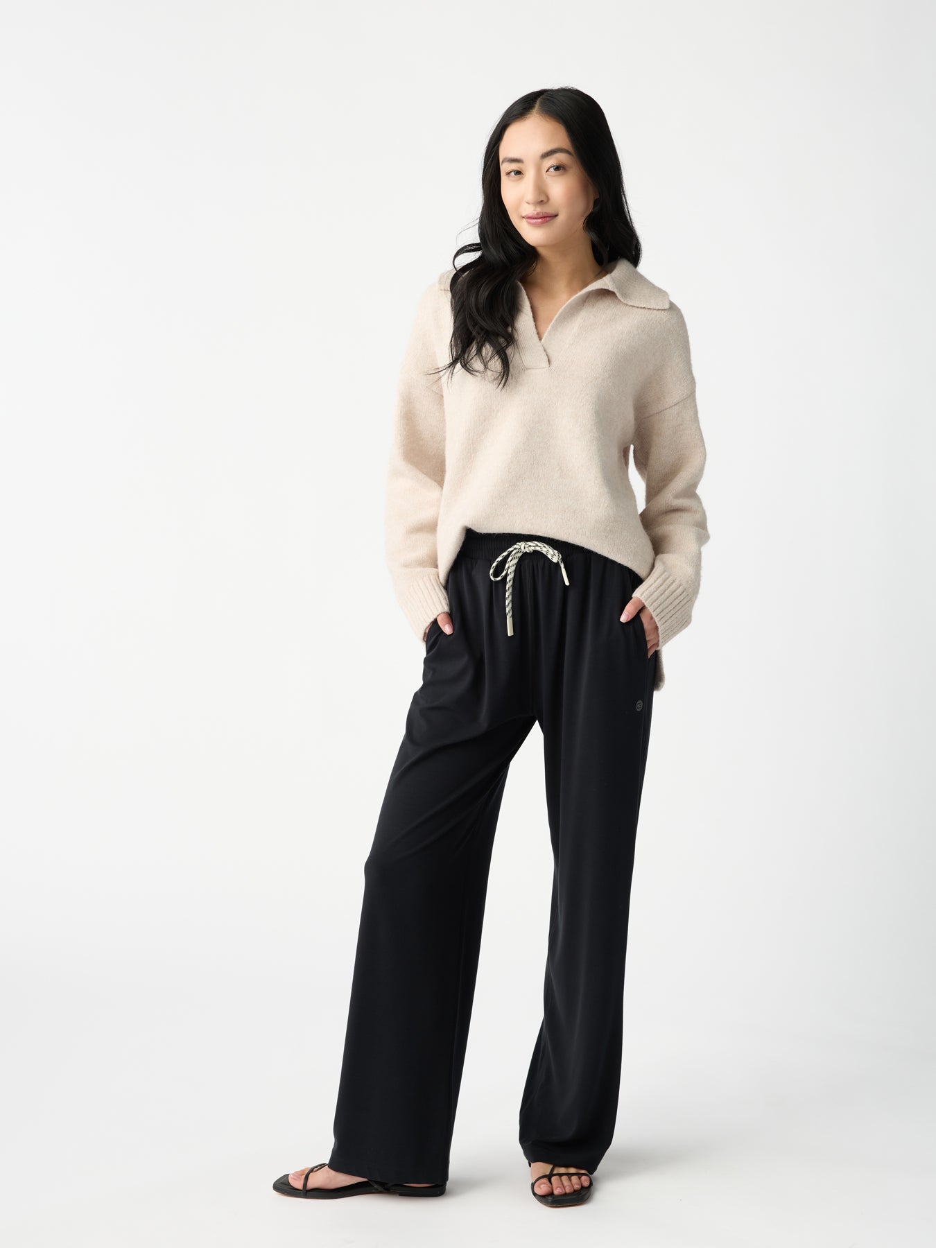 A person with long hair stands against a plain background, wearing a beige sweater with a half-zip neckline and Cozy Earth's Women's Studio Wide Leg Pant in black. Their hands are in their pockets, and they are wearing black sandals. 