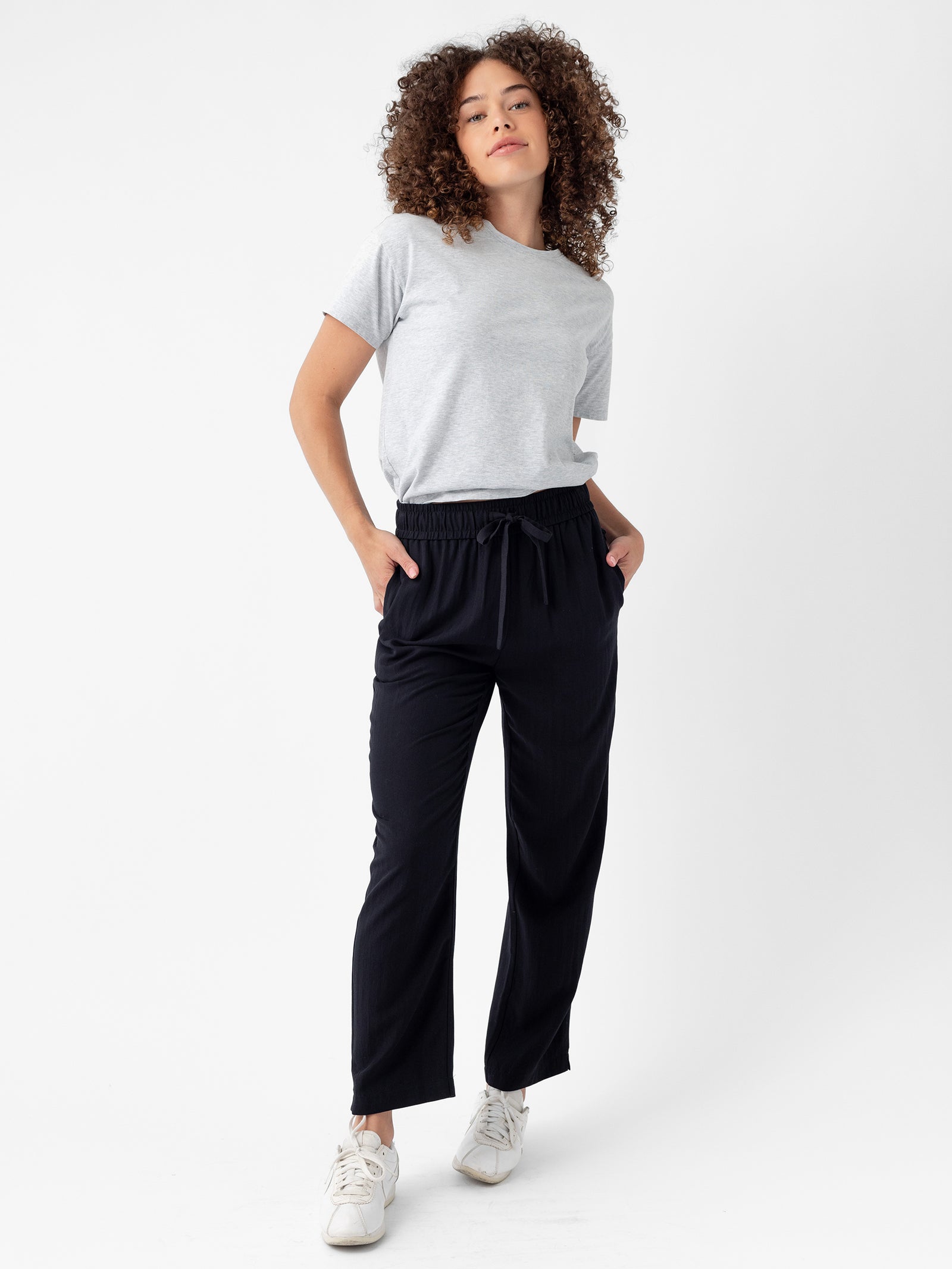 A person with curly hair is standing against a white background. They are wearing a light gray T-shirt, Cozy Earth's Women's Sunset Cropped Pants, and white sneakers. Their hands are in their pockets, and they have a relaxed expression on their face. 