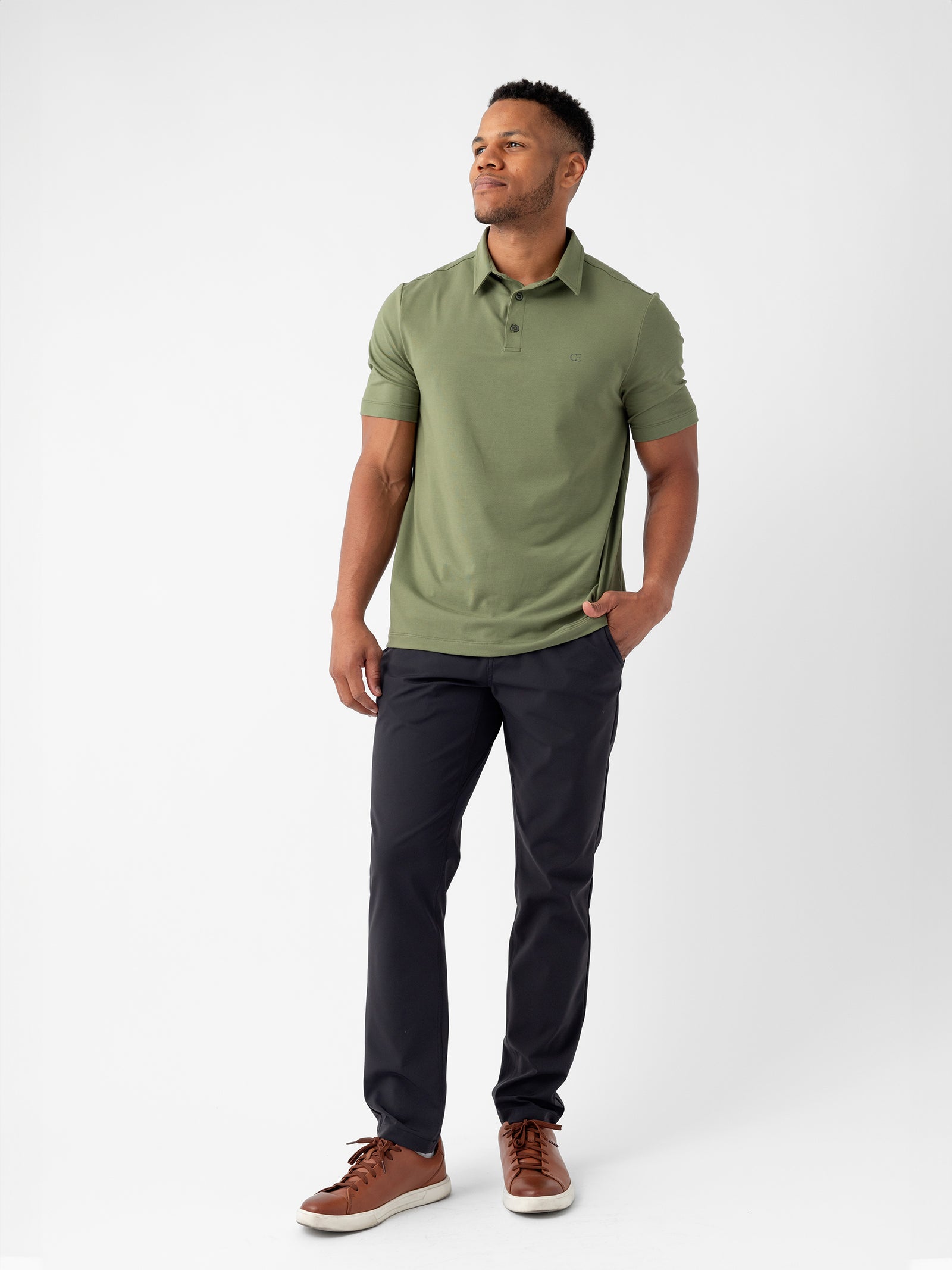 A man stands against a plain white background, wearing the Cozy Earth Men's Everyday Polo in green paired with dark pants and brown shoes. He has short hair, a slight beard, and is looking to the side with one hand in his pocket. 