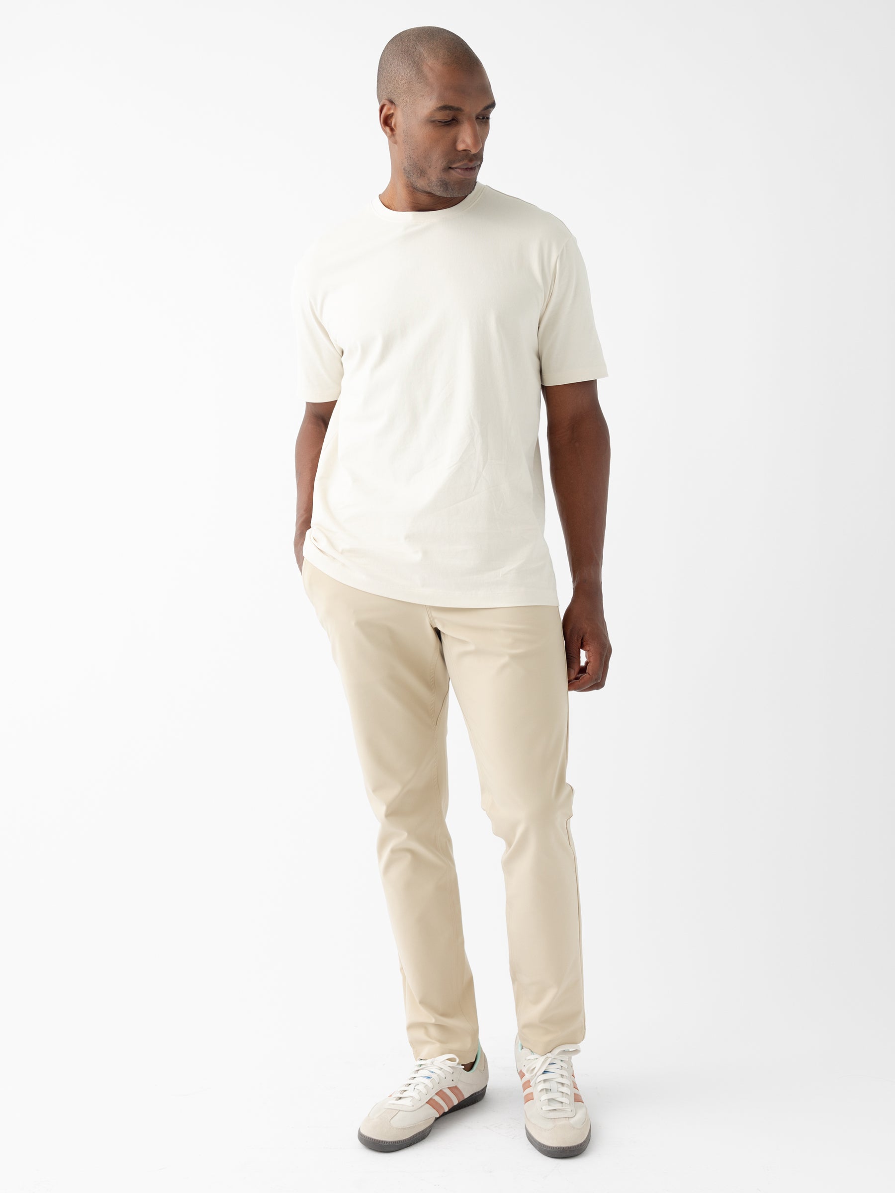 A person stands against a plain white background, looking down and to the side. They are wearing a casual cream-colored T-shirt, Cozy Earth's Men's Everywhere Pant 30L in beige, and white sneakers with gray and red detailing. The outfit is simple and monochromatic, emphasizing a clean and minimalistic style. |Color:Khaki