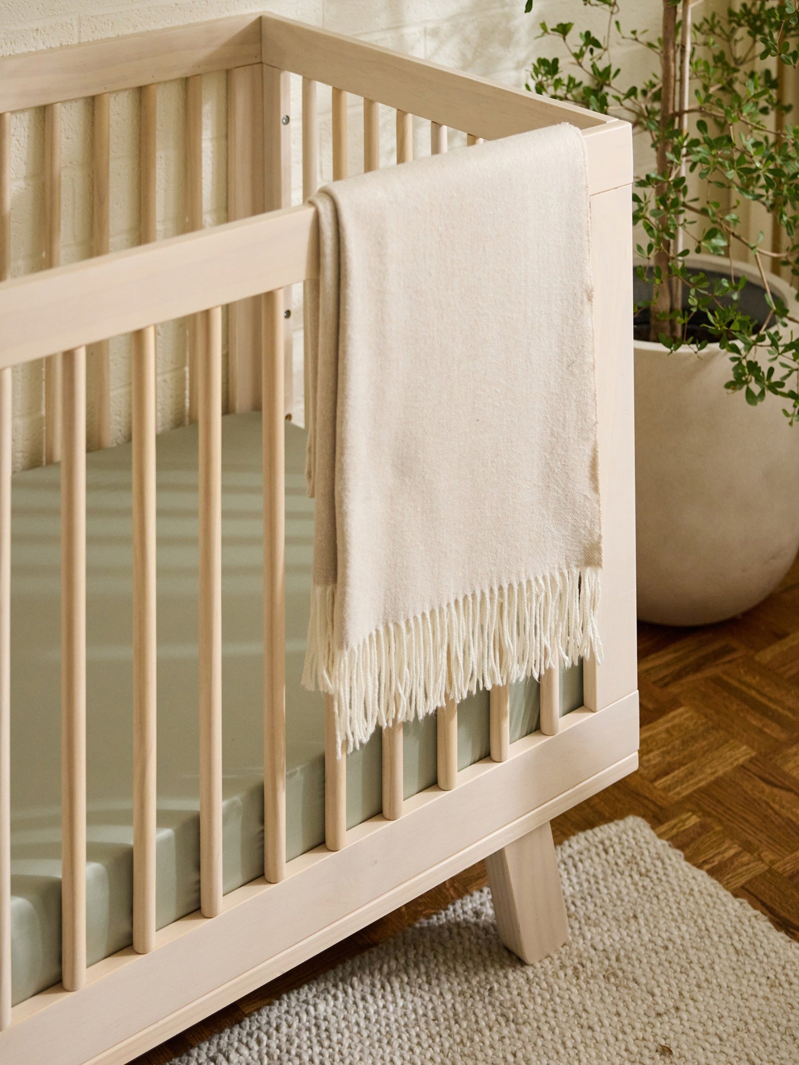 A light wooden crib with a Cozy Earth Crib Sheet, features a soft beige blanket draped over the side. Nearby, a potted plant sits on the woven rug partially visible on the wooden floor. 