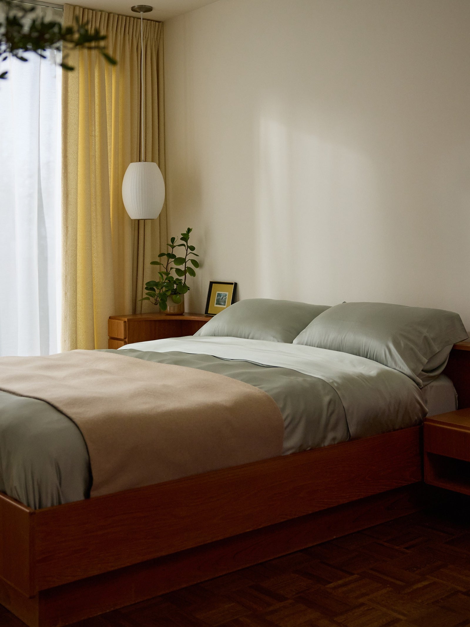 A minimalist bedroom with a wooden bed, Laurel Cozy Earth Bamboo Flat Sheet, and a beige throw. A small plant and framed picture sit on the bedside table. A white lamp hangs near opaque curtains, creating a calm, cozy ambiance. 