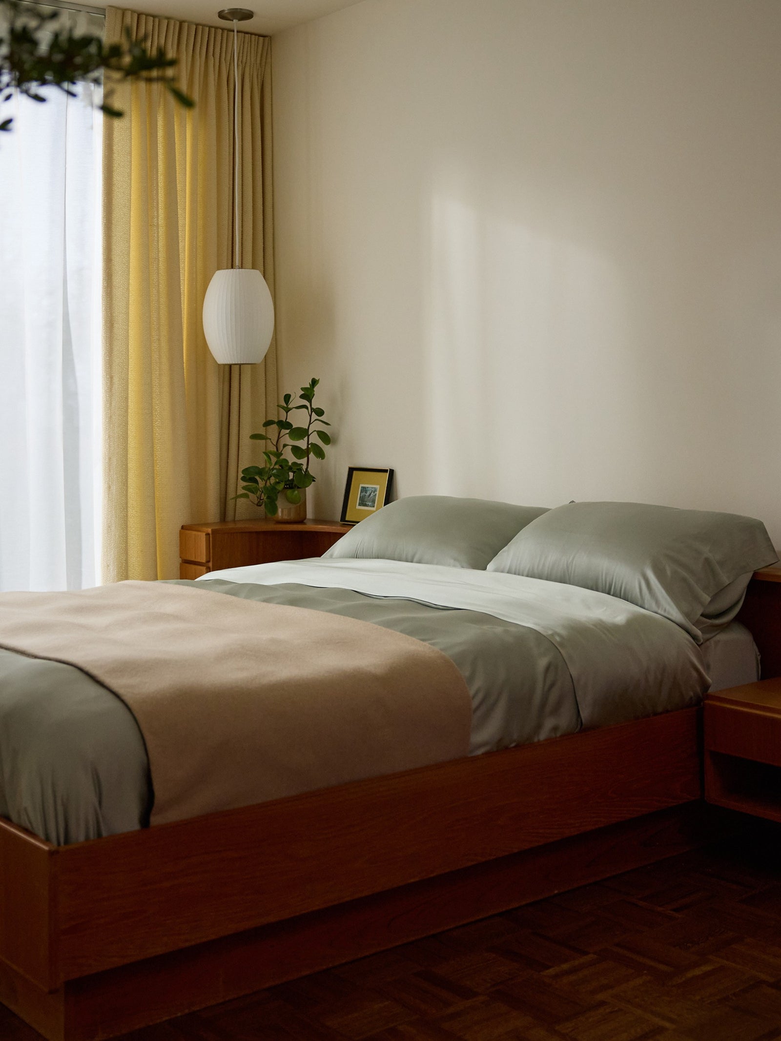 A calming bedroom features a wooden bed frame with neatly made green bedding, a beige blanket, and Cozy Earth's Bamboo Pillowcases. A small nightstand holds a plant and framed photo. Soft yellow curtains filter natural light through a large window, creating an inviting atmosphere. 