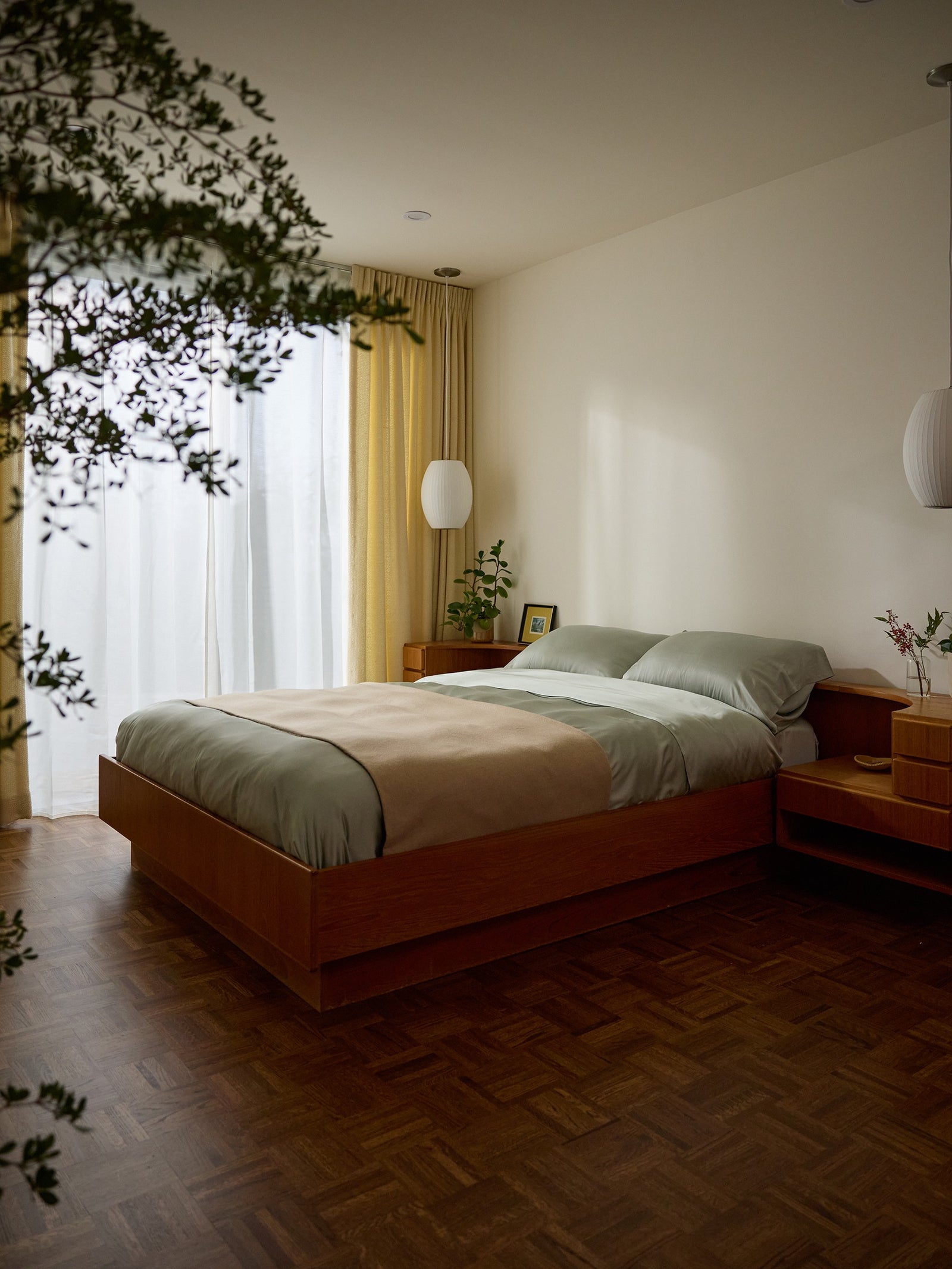 A cozy bedroom featuring a wooden bed frame adorned with Cozy Earth's Bamboo Sheet Set. Large windows with sheer and yellow curtains let in natural light, while a potted plant adds greenery and modern pendant lights hang beside the bed. 