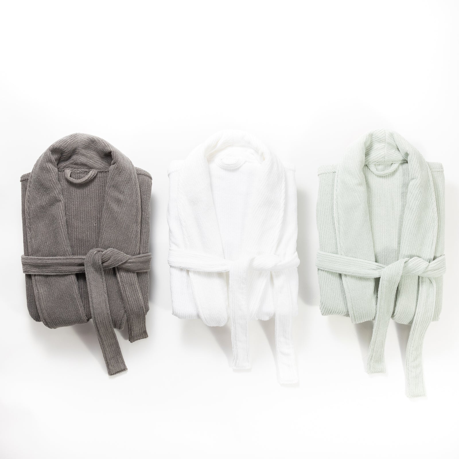 Three Ribbed Terry Bath Robes from Cozy Earth are folded and arranged side by side against a white background. From left to right, the robes are in dark gray, white, and light green colors. Each robe is neatly tied with a matching belt around the waist. 