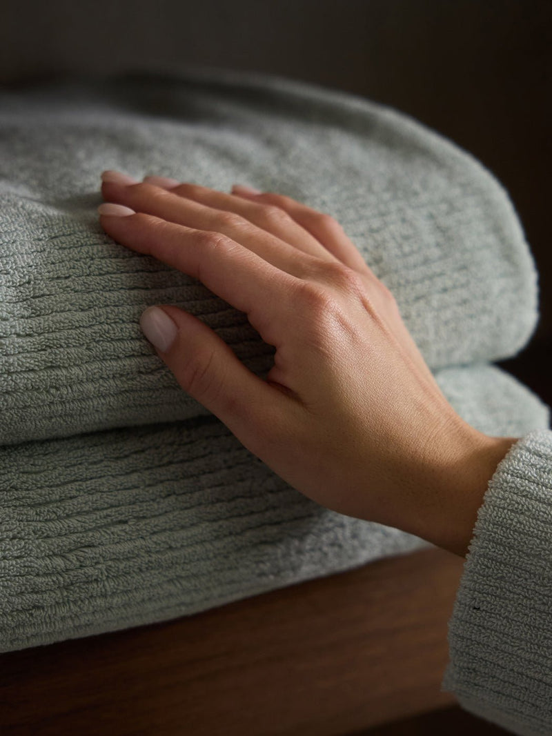 A hand gently rests on a neatly folded Ribbed Terry Bath Sheet by Cozy Earth, showcasing its textured gray fabric on a wooden surface.