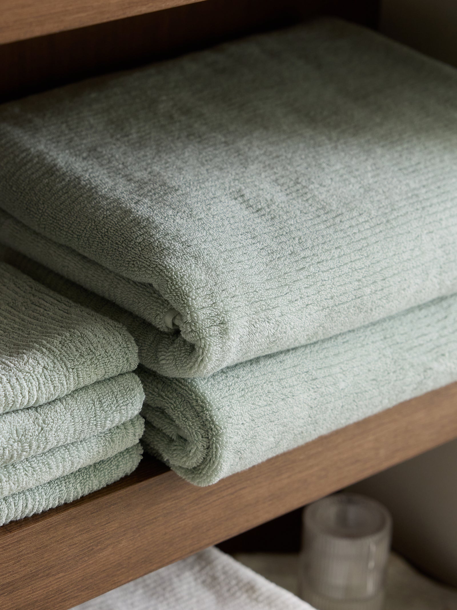 A stack of light grey Ribbed Terry Bath Towels by Cozy Earth is neatly arranged on a wooden shelf. In the background, another stack of similarly colored towels can be seen partially, along with a small white cylindrical candle holder placed underneath the shelf. 