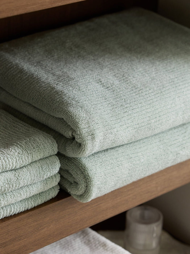 Two neatly folded Ribbed Terry Bath Towels by Cozy Earth in light green rest on a wooden shelf, with more towels and a frosted glass container nearby.
