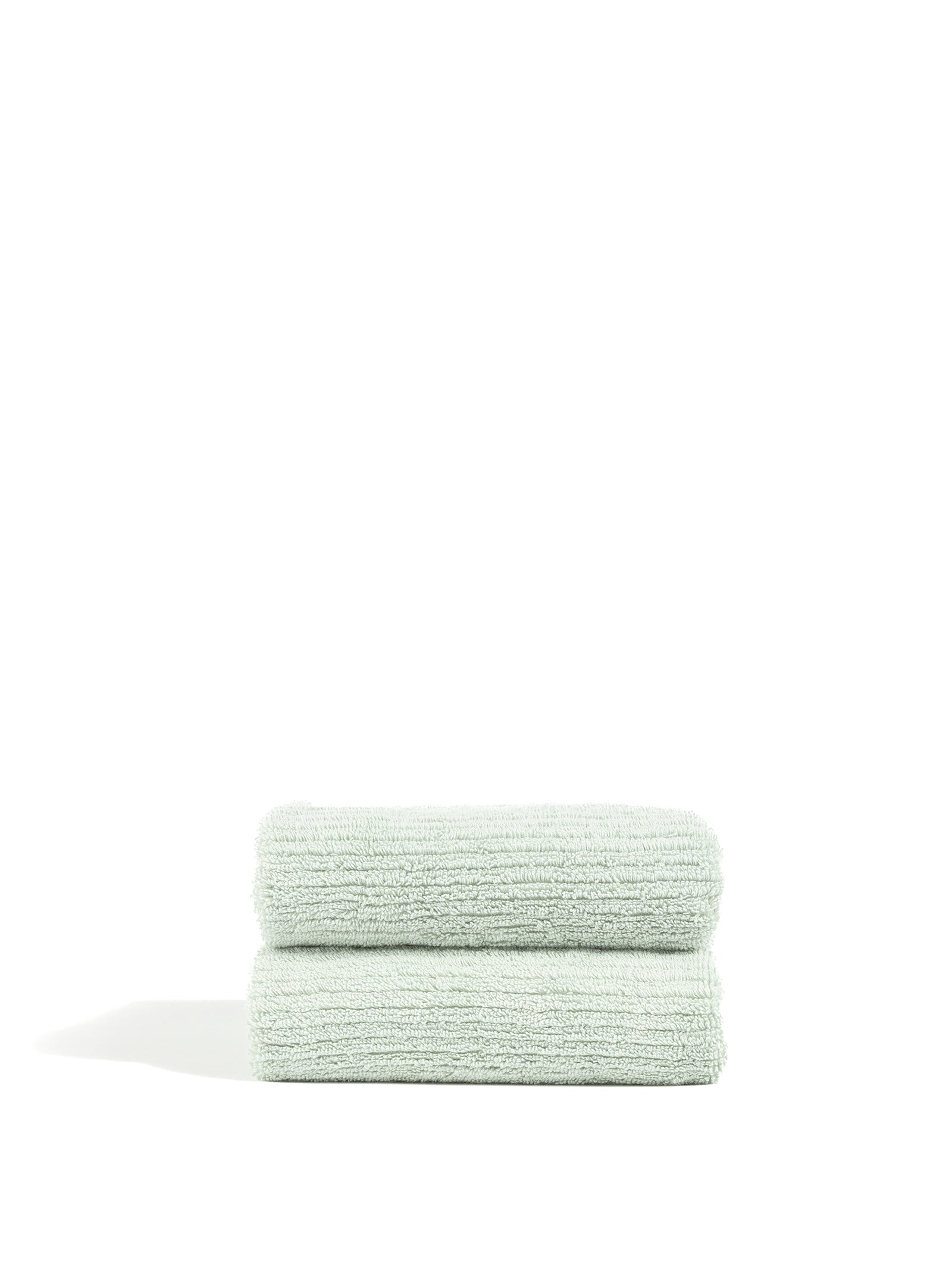 Two neatly folded Ribbed Terry Hand Towels from Cozy Earth, in a light green hue, are stacked on top of each other against a plain white background. The towels feature a soft, plush texture and boast a clean, minimalist appearance. 