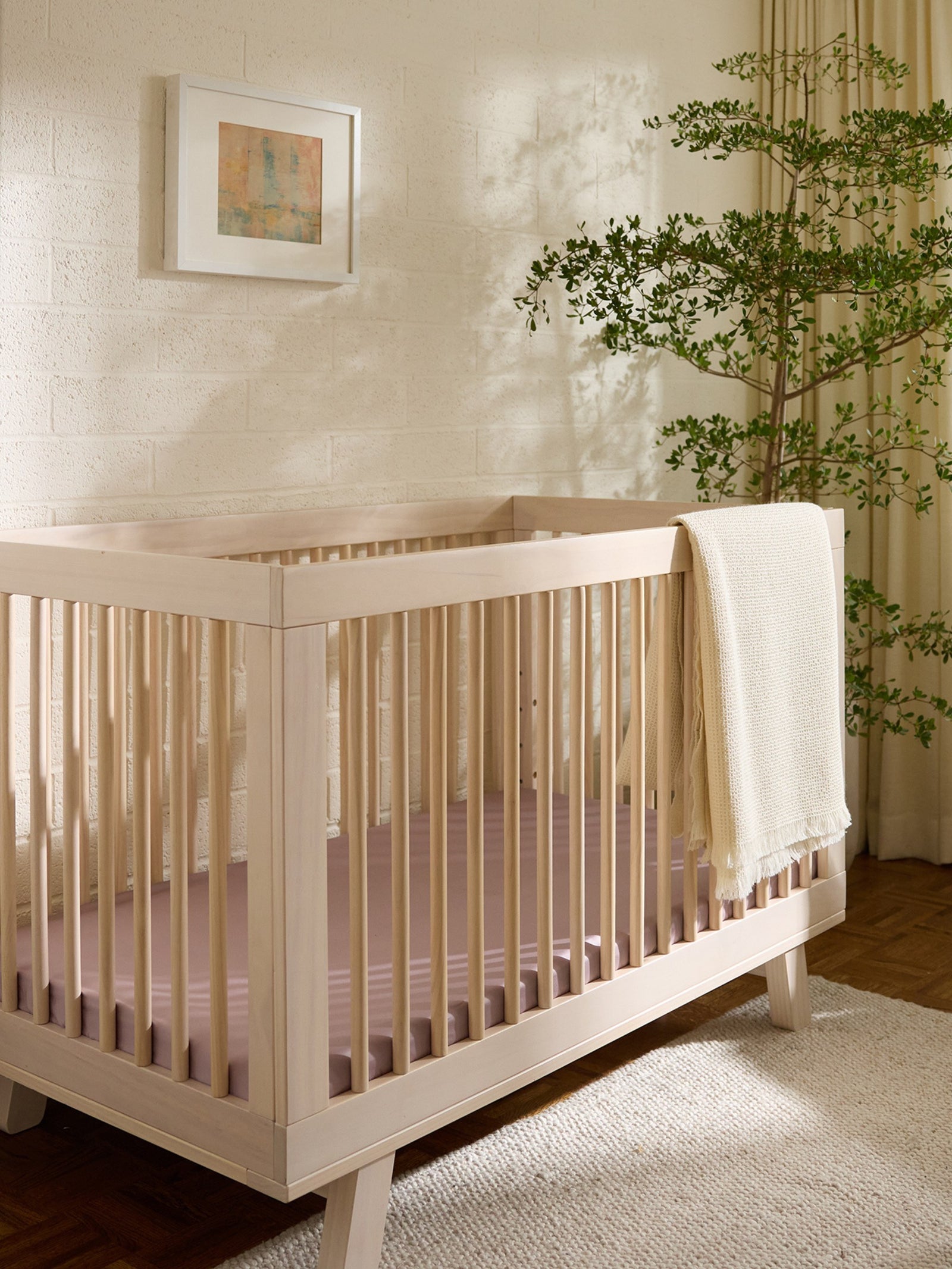 A minimalist nursery showcases a light wooden crib with a Cozy Earth Crib Sheet. The room includes a textured wall adorned with an abstract painting, a leafy plant in the corner, and a light-colored rug on the wooden floor. 