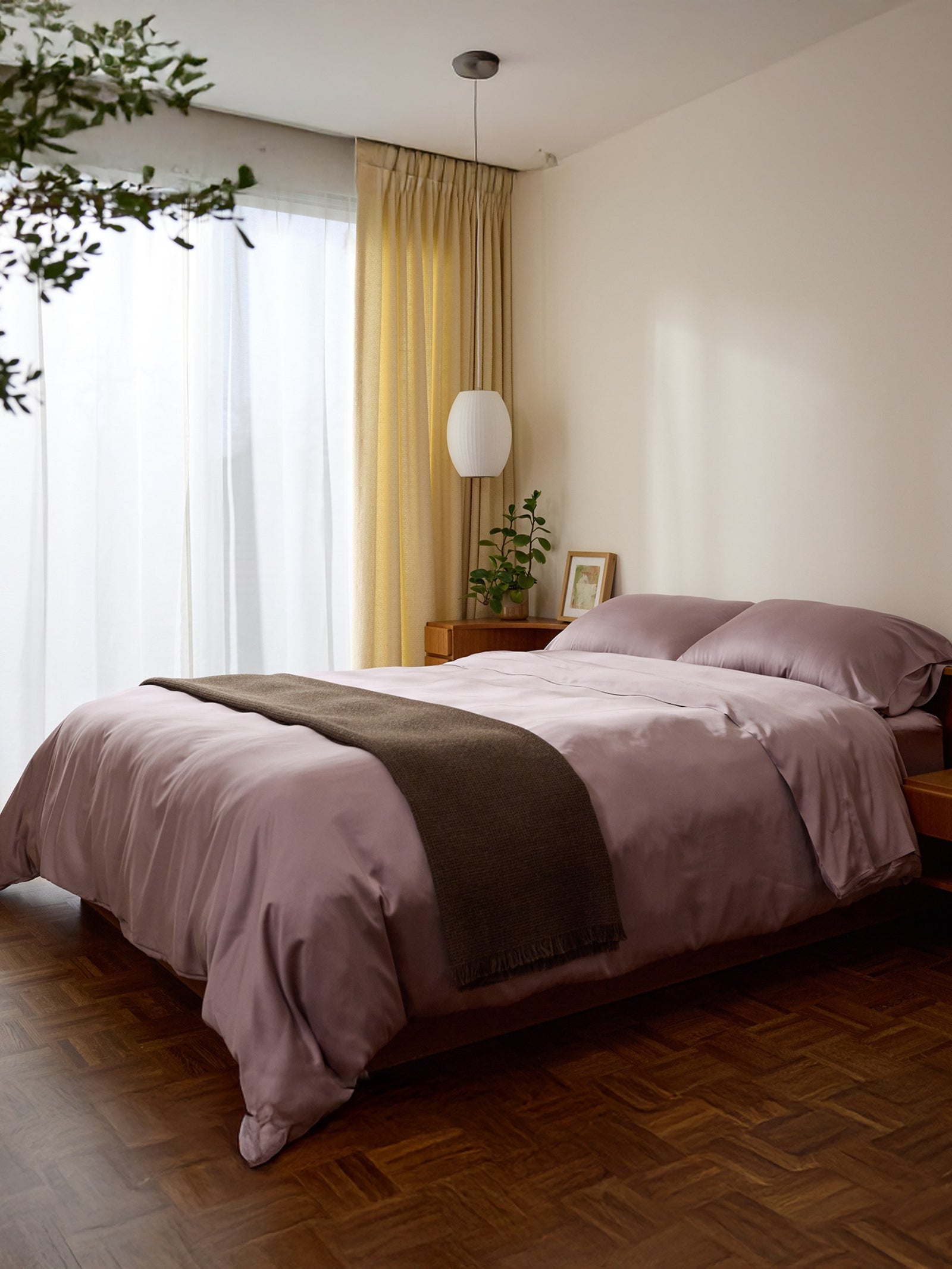 A cozy bedroom showcases a neatly made bed with Cozy Earth Bamboo Fitted Sheets in lavender, complemented by a dark gray throw. Soft light filters through sheer curtains, illuminating a potted plant on the wooden nightstand. A pendant light from the ceiling adds warmth to the space. 