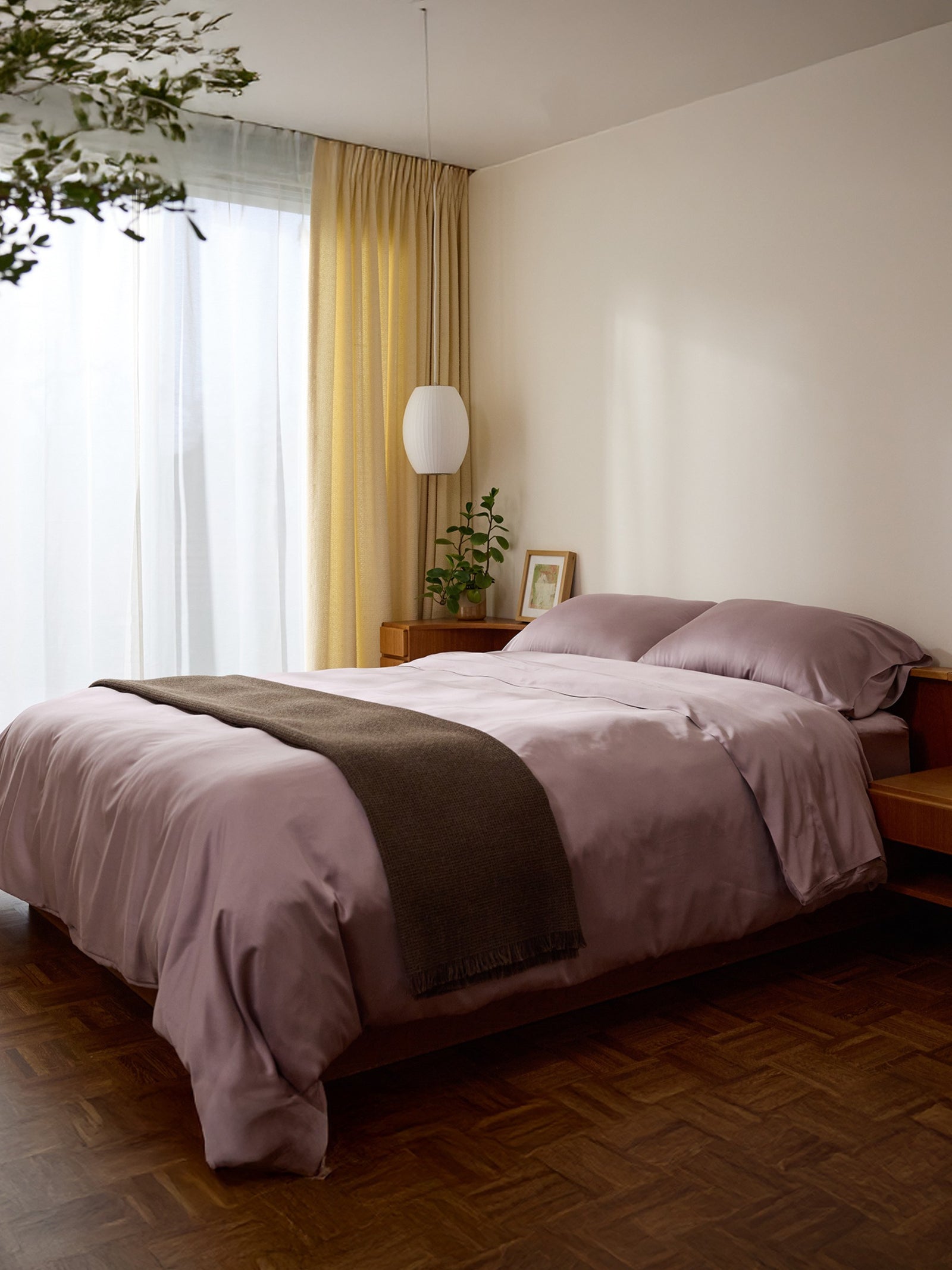 A cozy bedroom with a neatly made bed featuring Bamboo Pillowcases from Cozy Earth and a brown throw. Wooden furniture includes a bedside table with a plant and lamp. Yellow curtains hang beside a large window, allowing soft natural light to fill the room. 