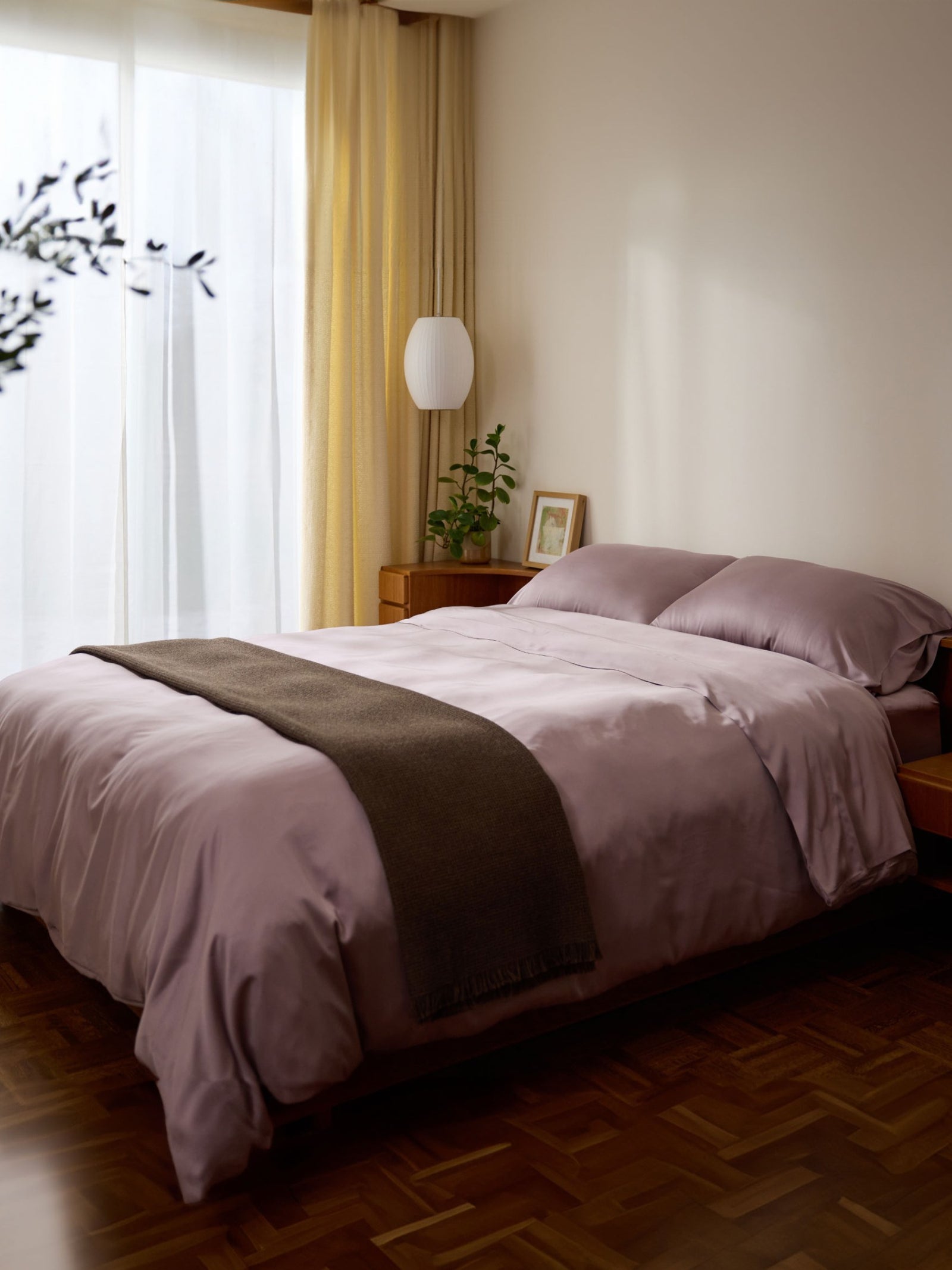 A cozy bedroom showcases a neatly made bed with Cozy Earth's Bamboo Sheet Set in light purple and a dark throw. A wooden nightstand holds a small green plant and framed photo, while soft light filters through sheer curtains to warm the room. 