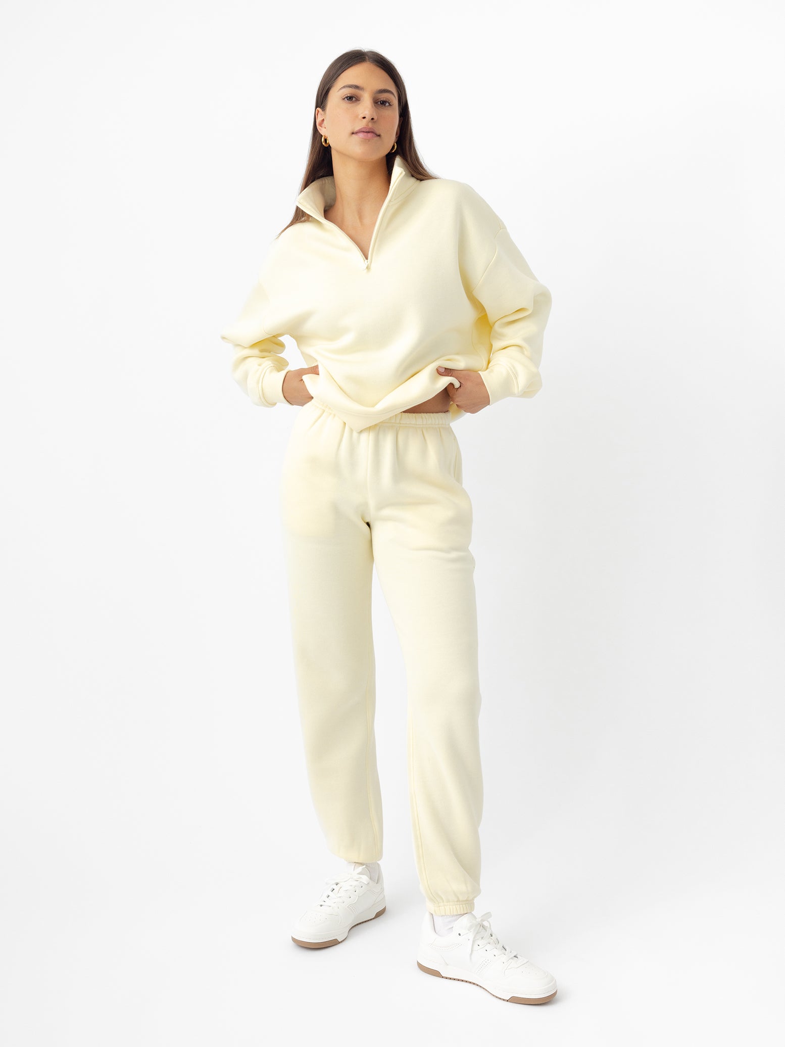 A person stands confidently in a light yellow Women's CityScape Sweatpant from Cozy Earth, featuring a zip-up top and matching joggers. They have long dark hair and white sneakers against a plain white background. |Color:Lemonade