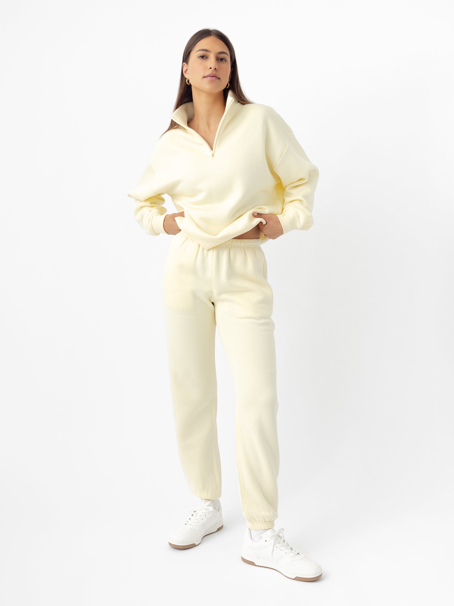 A person stands confidently in a light yellow Women's CityScape Sweatpant from Cozy Earth, featuring a zip-up top and matching joggers. They have long dark hair and white sneakers against a plain white background. 