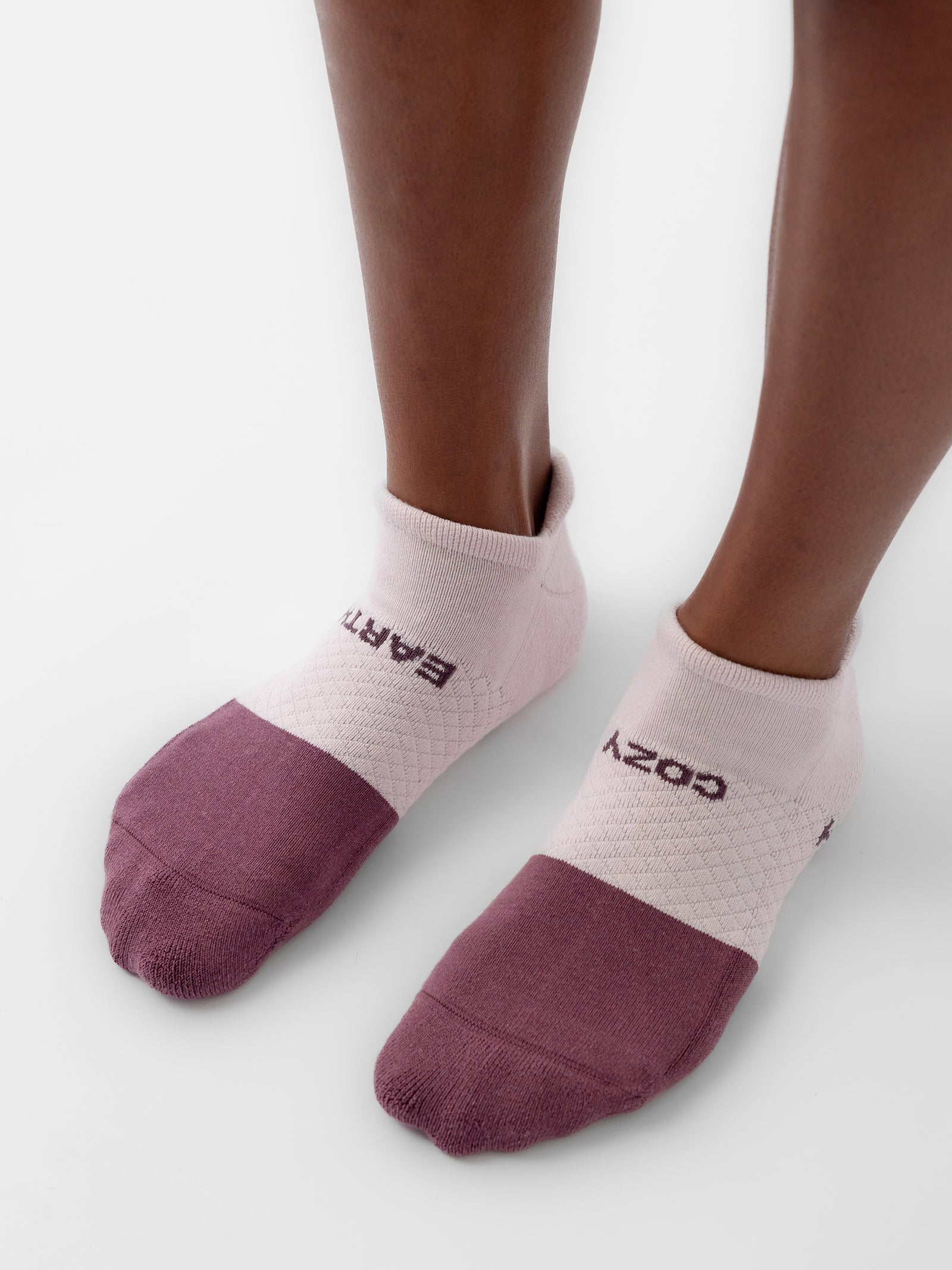 Close-up of a person's feet wearing Cozy Earth's Essential Ankle Sock 2-Pack. The socks feature purple toe and heel sections, a white middle section with a textured pattern, and "COZY EARTH" printed on the top. The background is plain white. 