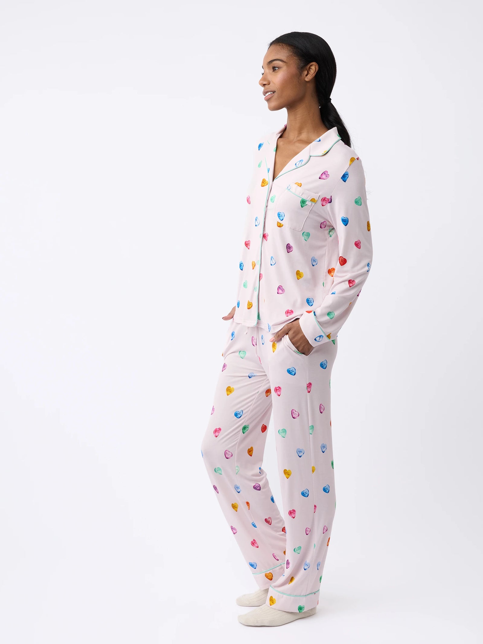 A person stands sideways wearing the Women's Stretch-Knit Bamboo Pajama Pant by Cozy Earth, featuring colorful heart patterns on a light background. They have their hands in the pockets and are smiling against a plain white backdrop. 