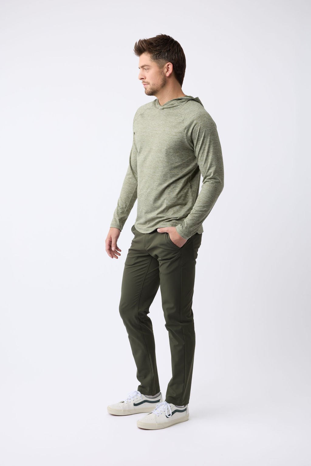 A man in a Cozy Earth Men's Lightweight Performance Hoodie, light green with long sleeves, paired with dark green pants and white sneakers. His short hair complements the look as he stands against a plain white background, gazing sideways. 