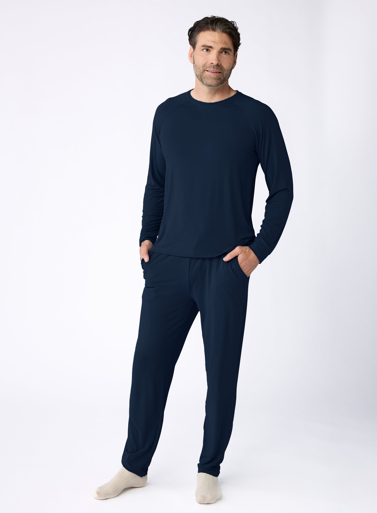 A man wears the Cozy Earth Men's Bamboo Stretch-Knit Long Sleeve Pajama Set in dark blue, hands in pockets and paired with beige socks against a plain white background.