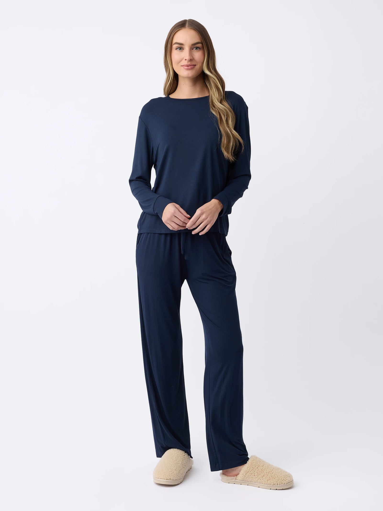 A woman with long hair wears the Women's Stretch Knit Bamboo Long Sleeve Lounge Tee by Cozy Earth in navy blue, paired with matching pants and beige slippers. She stands against a plain white background, looking relaxed and comfortable. 