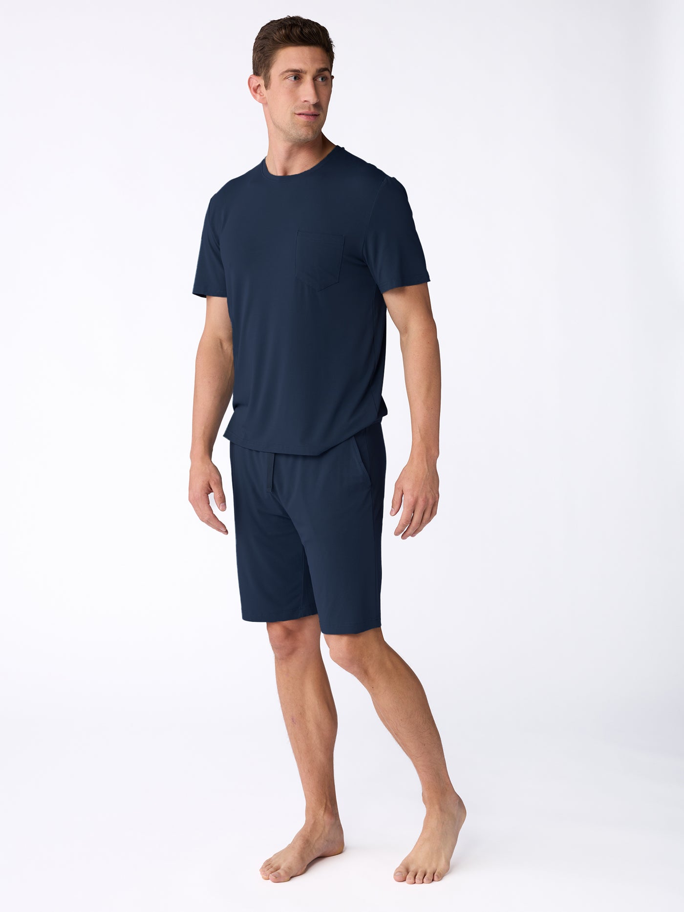 A man stands barefoot against a white background, wearing the Cozy Earth Men's Bamboo Stretch-Knit Short Sleeve Pajama Set in navy blue. He looks to his left with a relaxed posture, showcasing the set's casual and comfortable style. 