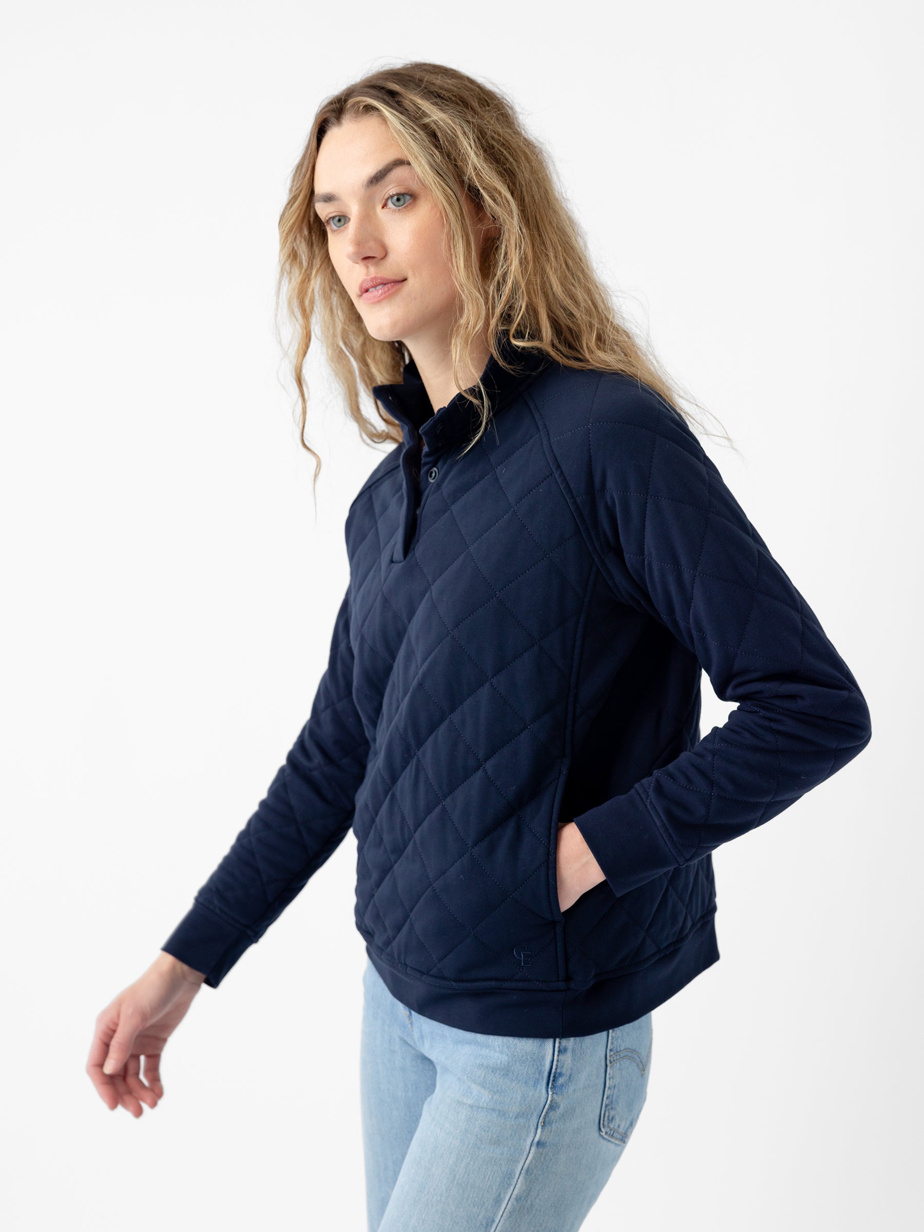 A person with long, wavy hair wearing the Cozy Earth Women's Ultra-Soft Bamboo Quilted Snap Pullover in dark blue and light blue jeans stands against a plain white background. They are looking slightly off to the side with one hand in a pullover pocket. |Color:Navy