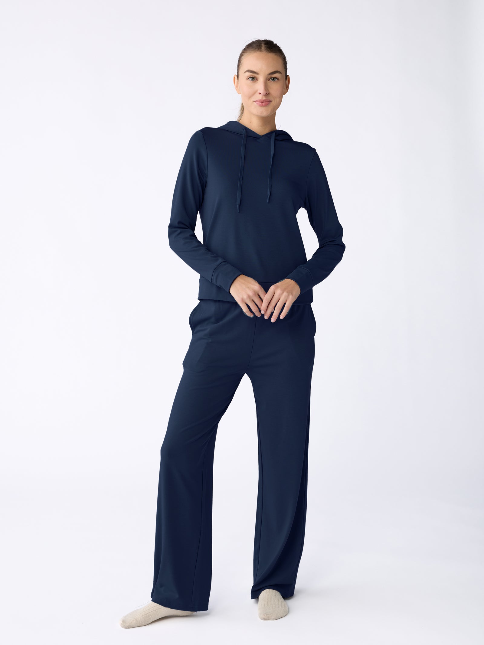 A person stands against a plain backdrop in the dark blue Women's Ultra-Soft Bamboo Wide Leg Pull On Pant & Hoodie Set from Cozy Earth, wearing light-colored socks, facing forward with hands clasped. 