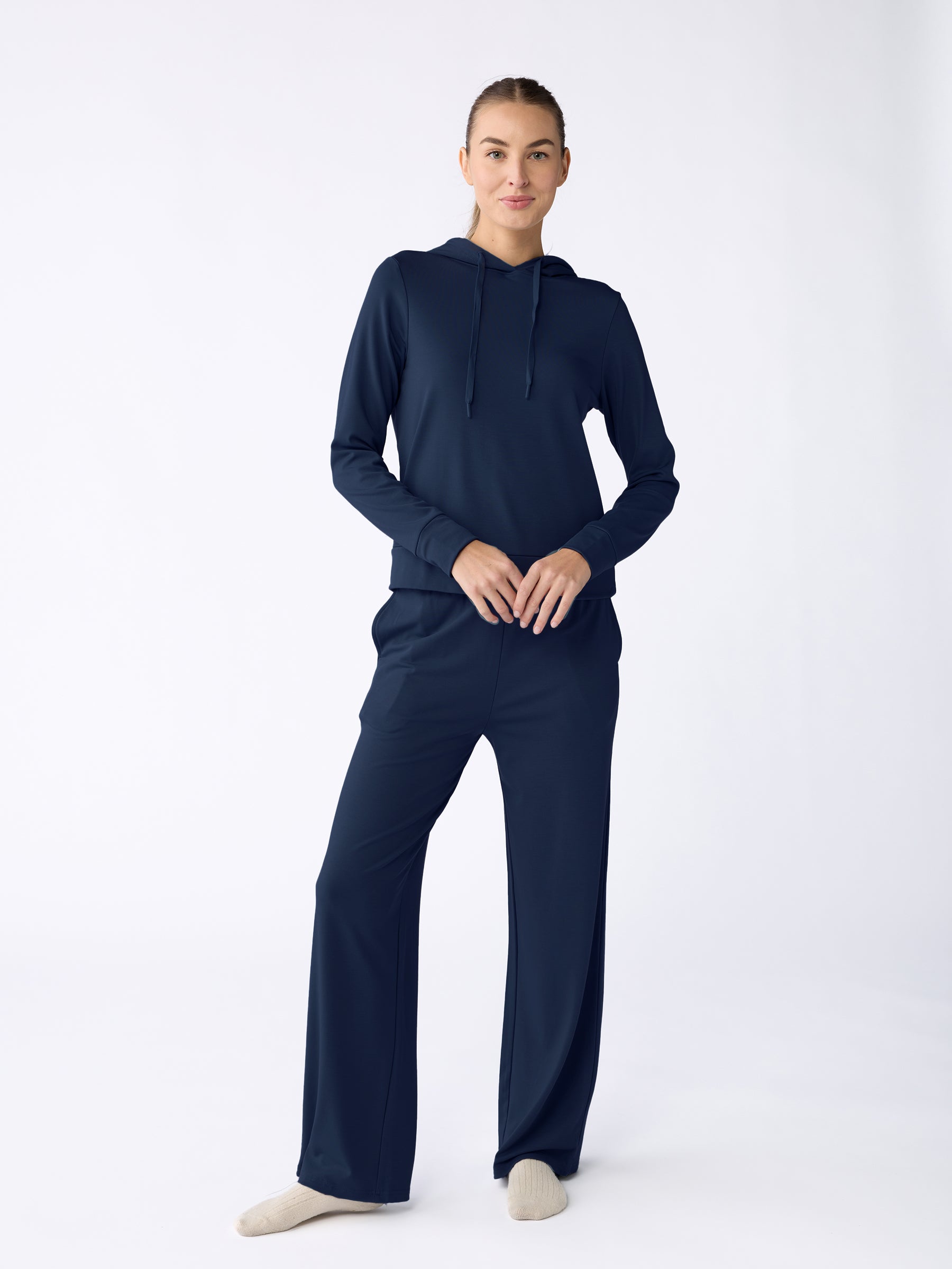 A person stands against a plain backdrop in the dark blue Women's Ultra-Soft Bamboo Wide Leg Pull On Pant & Hoodie Set from Cozy Earth, wearing light-colored socks, facing forward with hands clasped. |Color:Navy