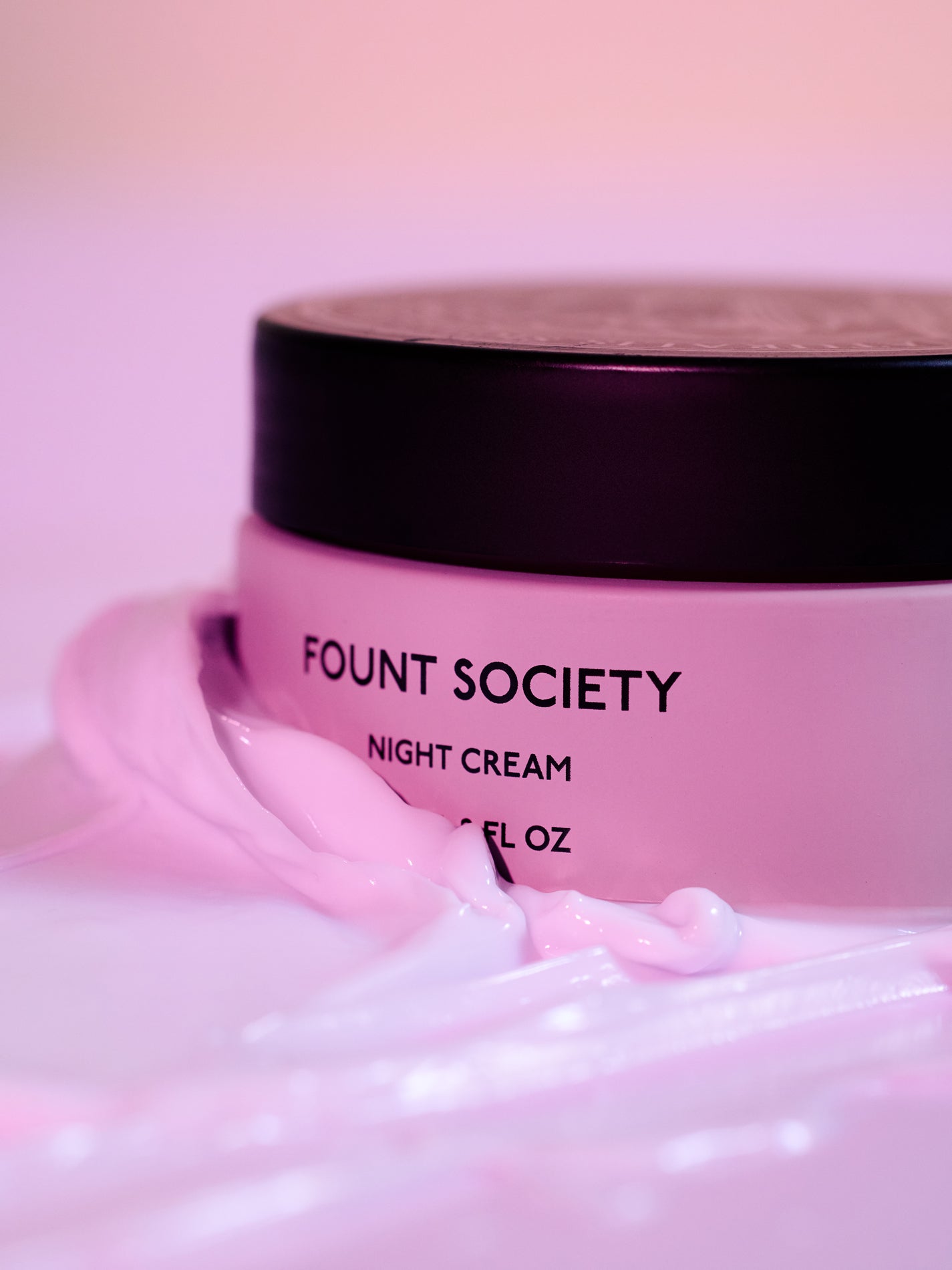 A pink jar of Cozy Earth Night Cream with a black lid, surrounded by swirls of light pink cream, sits against a softly blurred pink background. 