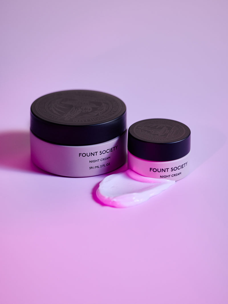 Two jars of Cozy Earth Night Cream rest on a pink surface. The larger jar is partially open, revealing a creamy smear beside the smaller, closed jar. Both have black lids.