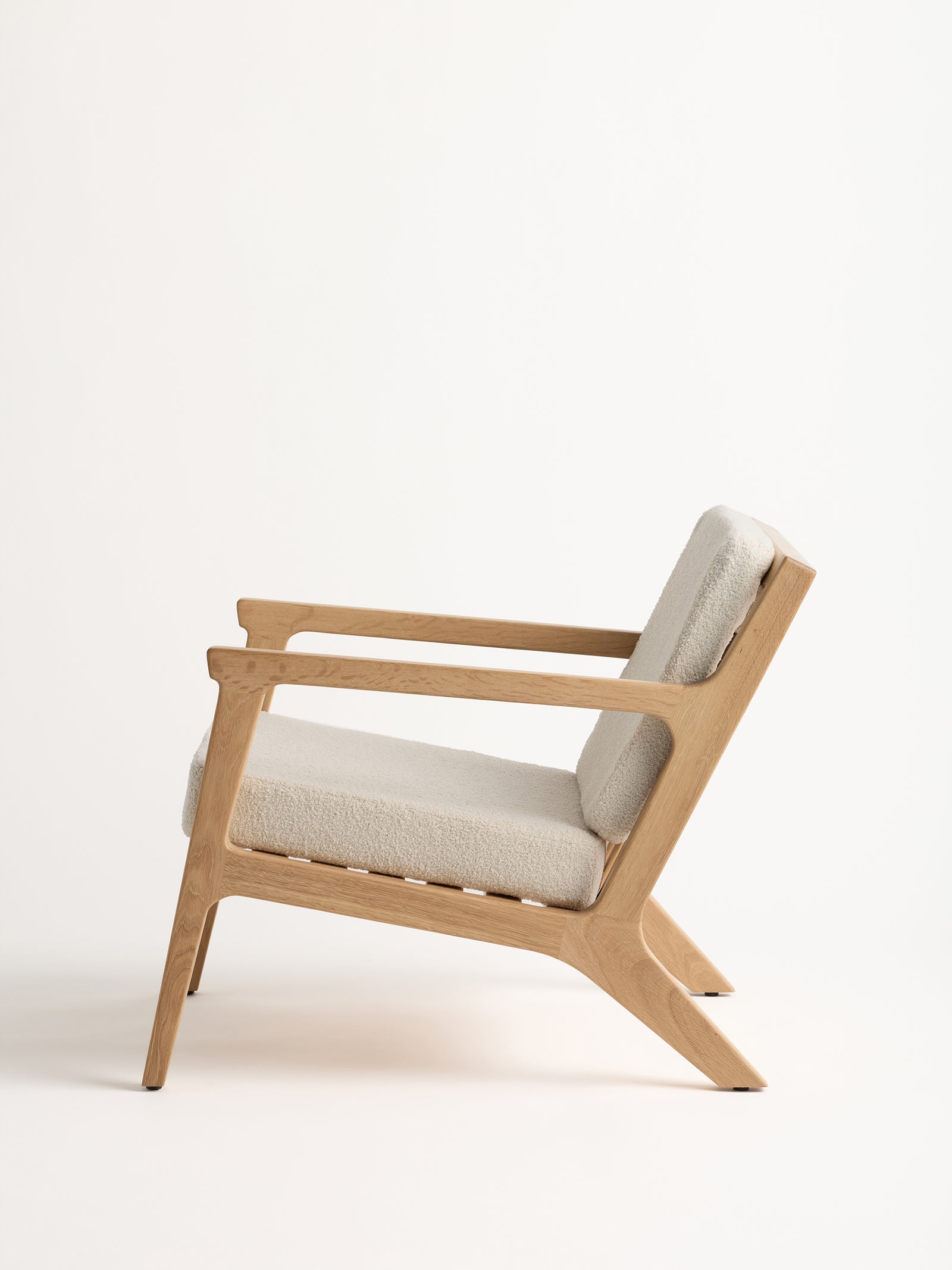 The Coronado Lounge Chair by Cozy Earth is depicted in a side profile view against a plain white background. This wooden chair boasts a minimalist design, featuring a light beige cushioned seat and backrest, angled legs, and open armrests. 