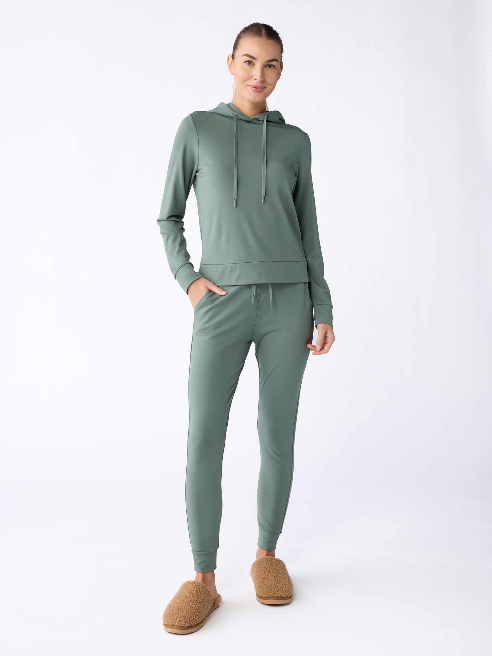 A person stands against a light background, dressed in a matching sage green set from Cozy Earth's Women's Ultra-Soft Bamboo Hoodie collection, with tan slippers. Their hair is styled in a neat bun, hands tucked into pockets, and they are smiling. 