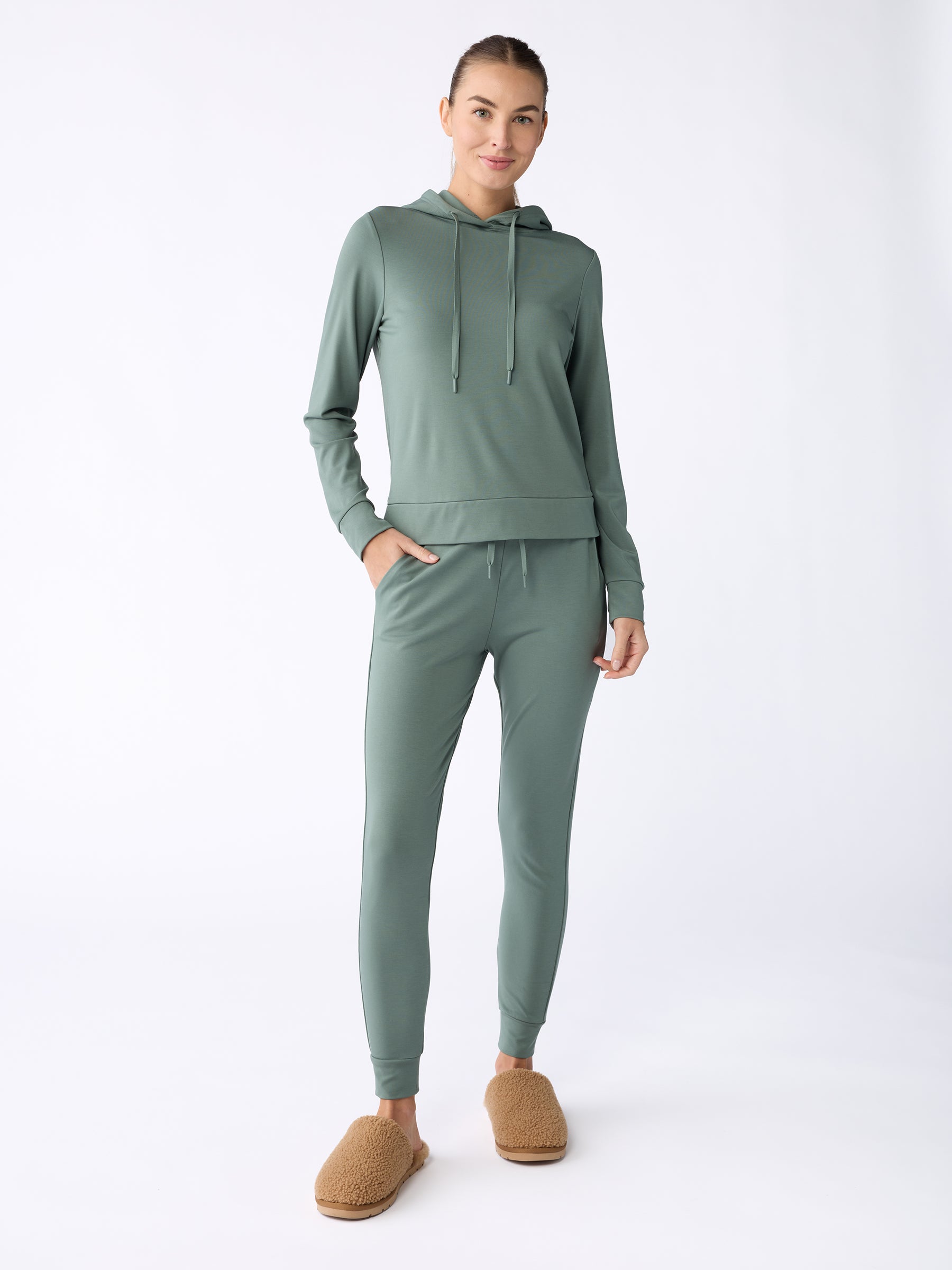 A person dressed in a light green hoodie paired with Cozy Earth's Women's Bamboo Jogger Pant stands against a plain background. They are also wearing brown fuzzy slippers, have their hair tied back, and are smiling slightly. |Color:Oakmoss