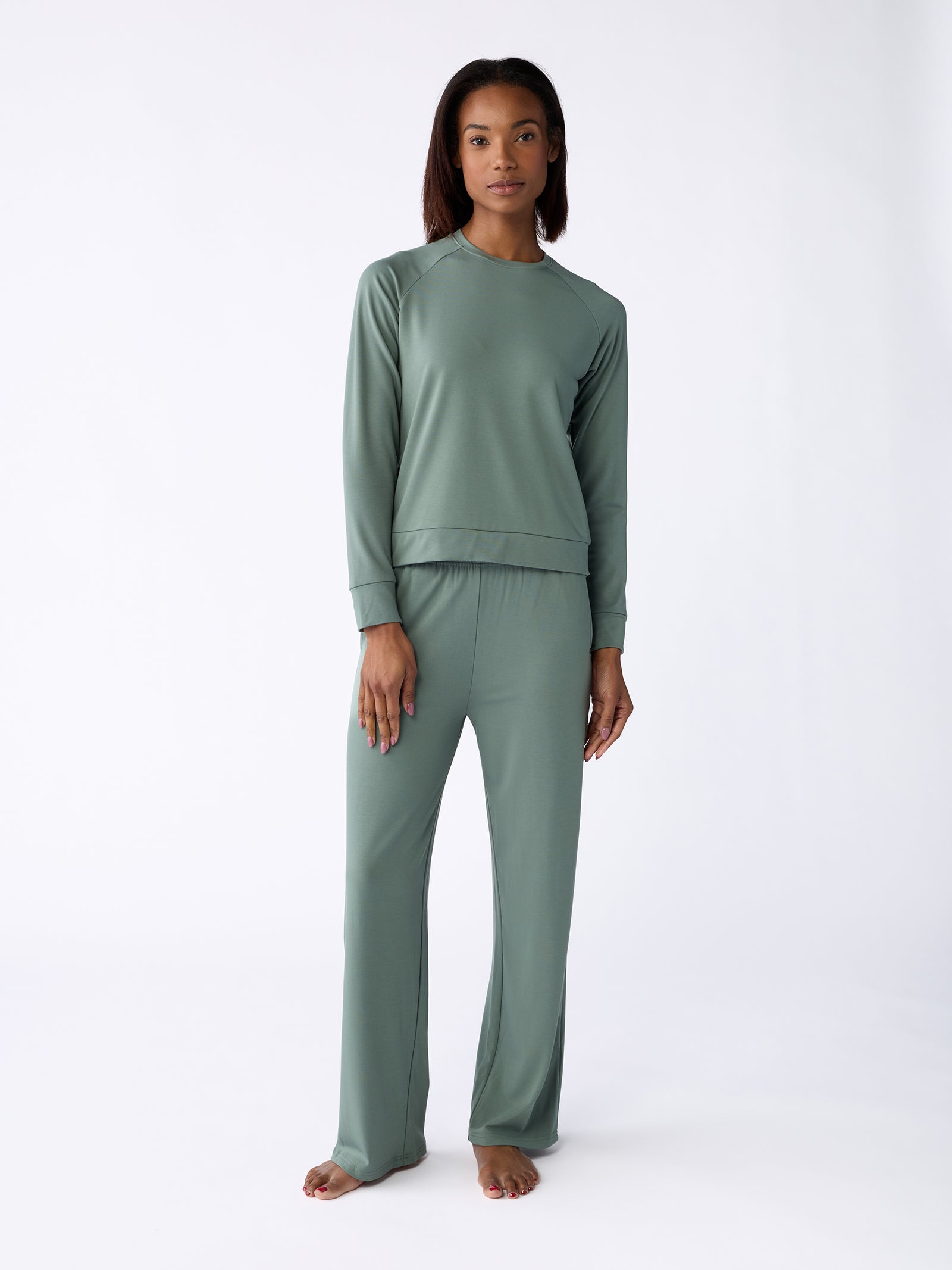 A woman with long dark hair stands barefoot on a white background, wearing the Cozy Earth Women's Ultra-Soft Bamboo Pullover Crew in sage green, paired with matching pants, as she looks directly at the camera. 