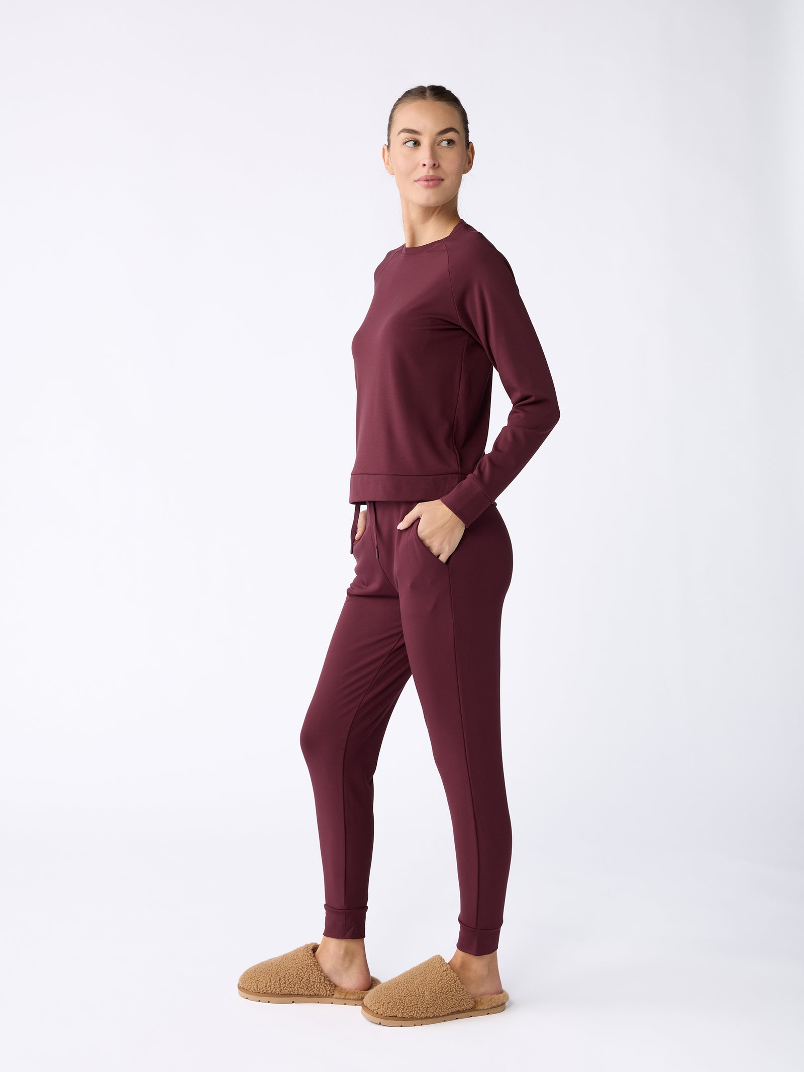 A person stands against a white background, wearing the Women's Ultra-Soft Bamboo Pullover Crew by Cozy Earth in dark red, paired with matching lounge pants and brown slippers. They have their hands in the pockets of their pants and are looking slightly to the side. 