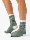 A person wears Cozy Earth’s The Fair Isle Plush Lounge Sock, a light green style with white toes, heels, and cuffs, plus black-and-white geometric patterns on the cuffs and near the toes. The background is white.
